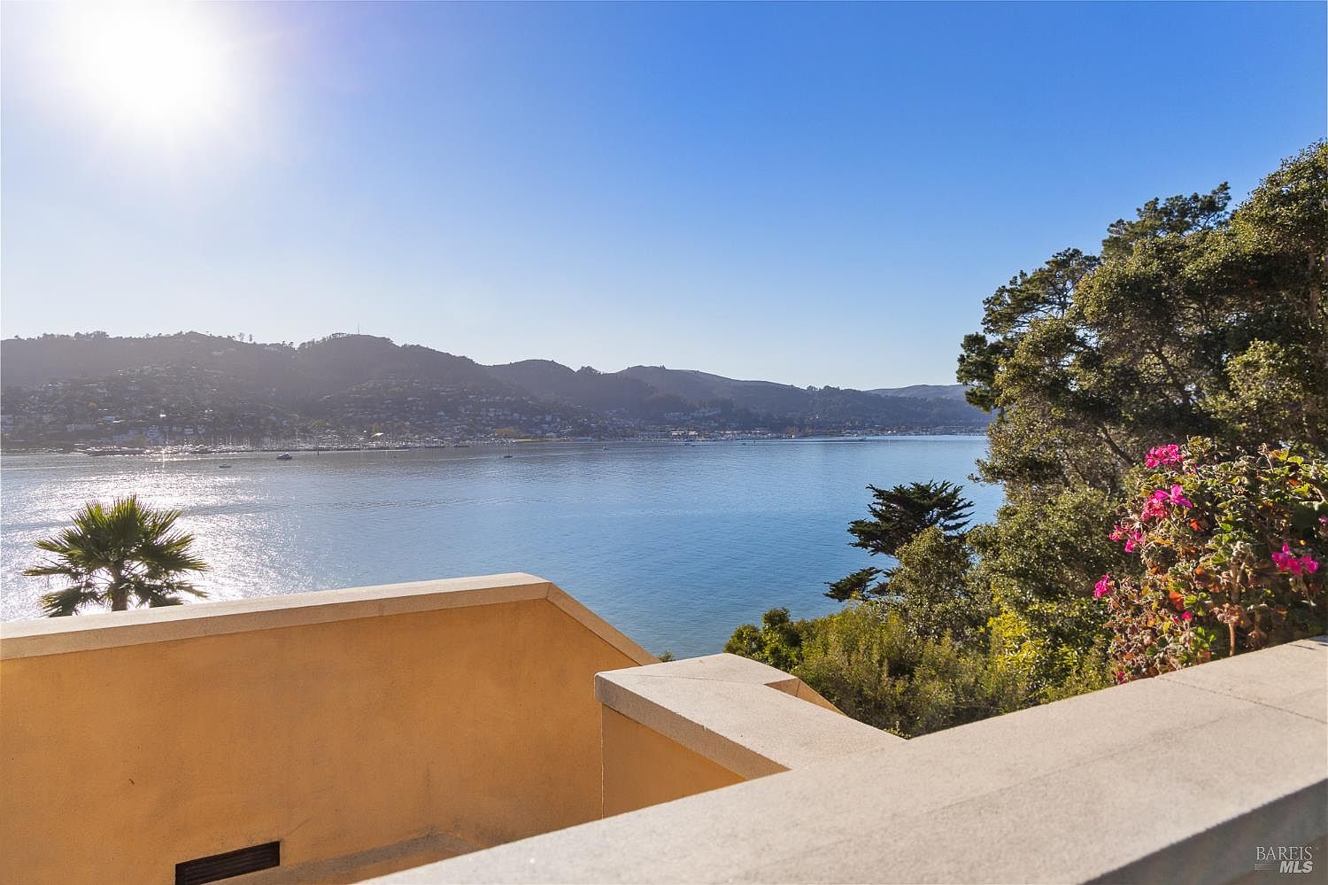 This elevated balcony offers a panoramic view of a serene bay, framed by lush green trees and distant hills. The foreground features a tan-colored stucco railing and a glimpse of a staircase, creating a sense of height and privacy. The bright, sunny atmosphere highlights the tranquil waterfront setting, making it an ideal space for outdoor relaxation.