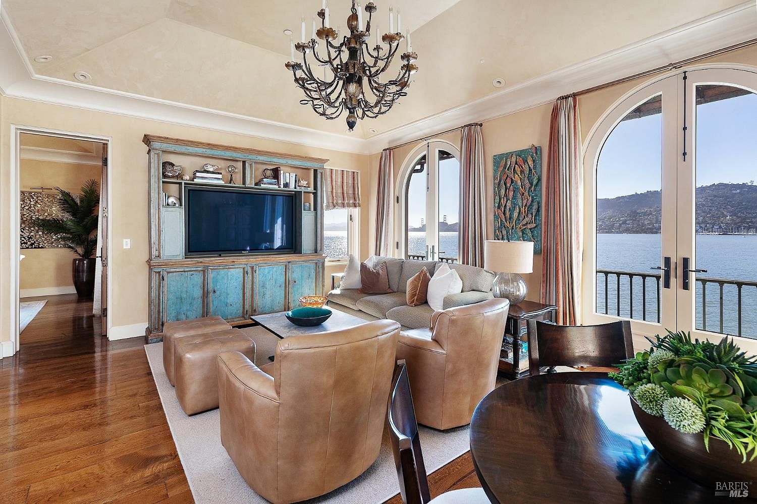 This elegant living room features a sophisticated coastal aesthetic with a prominent blue-distressed media cabinet and a grand chandelier as the centerpiece. The space is furnished with comfortable leather armchairs and a plush sofa, all oriented toward a scenic view of the water through large arched windows. The warm wood flooring and neutral color palette create an inviting atmosphere, while the open layout seamlessly connects to an adjacent dining area.