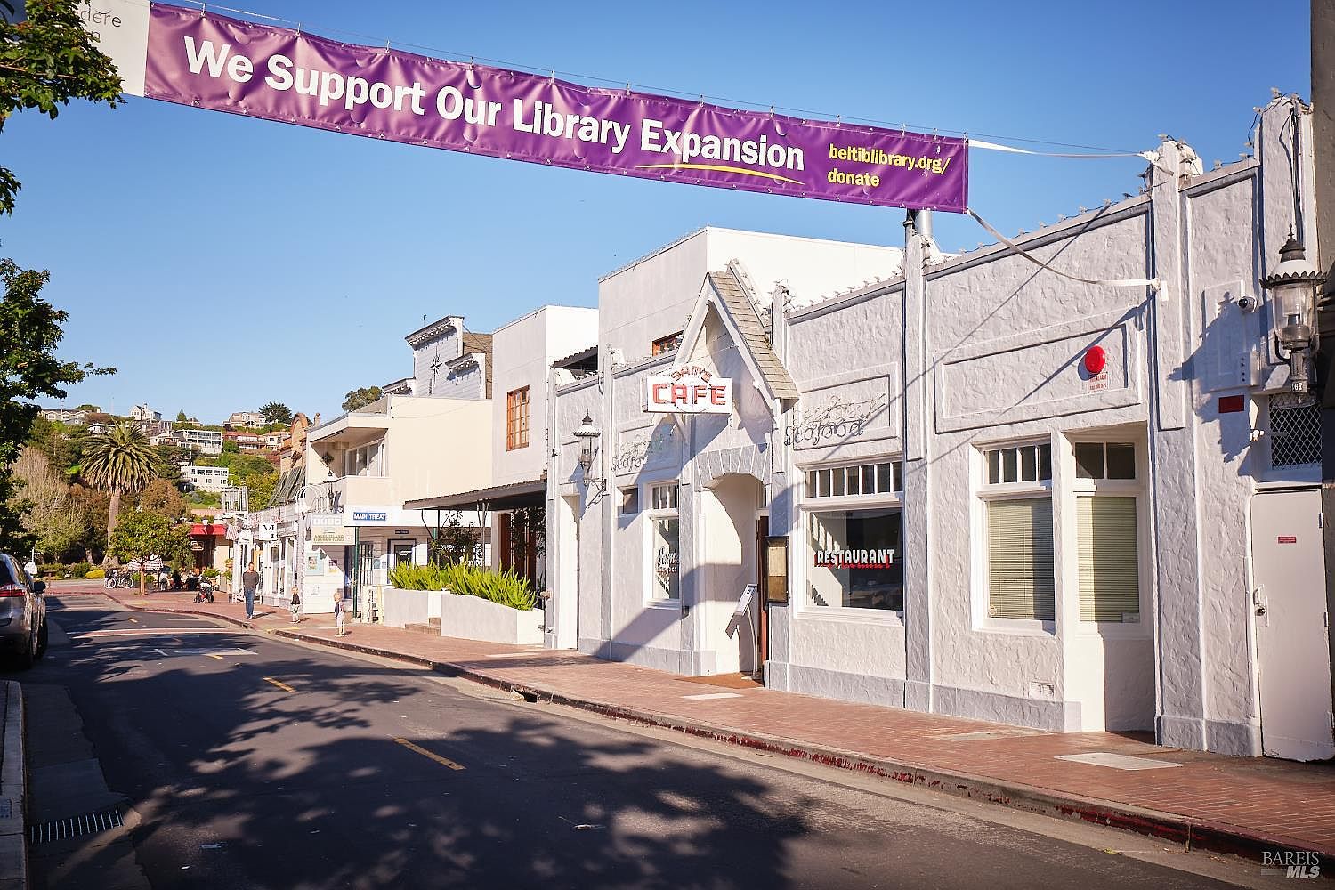 This street-level view captures a charming commercial block featuring white stucco buildings with classic architectural details, including a prominent 'Sam's Cafe' sign. A large purple banner spans the street, promoting a library expansion, while the scene is bathed in warm, late-afternoon sunlight. The perspective looks down a quiet, tree-lined street with a clear view of the storefronts and the hillside residential area in the background.