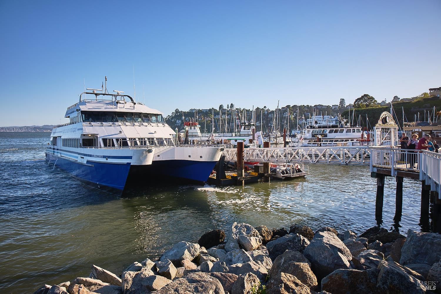 This scenic waterfront view captures a large passenger ferry docked at a marina, with a wooden pier extending toward the right side of the frame. The foreground features a rocky shoreline, while the background showcases a hillside community with numerous boats moored in the harbor under a clear blue sky. The perspective is a wide-angle, eye-level shot that emphasizes the active maritime lifestyle and coastal setting of the property's location.
