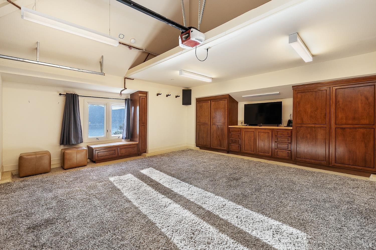 This finished garage space features a carpeted floor with a unique white-striped design, suggesting a multi-purpose recreational area. The room is equipped with built-in dark wood cabinetry, a television, and a window seat overlooking a water view, blending utility with comfort. Overhead lighting and a garage door opener mechanism are visible on the ceiling, highlighting the functional nature of this converted space.