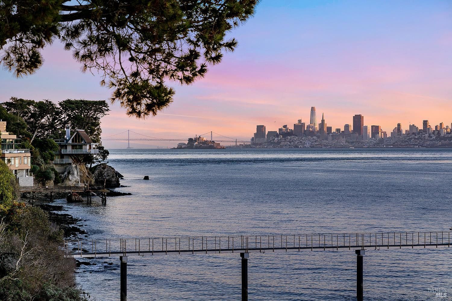 This scenic exterior view captures a waterfront property perched on a rocky cliffside, overlooking the San Francisco Bay. The composition features a multi-level home integrated into the landscape, with a long pier extending into the water and a panoramic backdrop of the city skyline and the Bay Bridge at sunset. The perspective is cinematic, emphasizing the dramatic coastal setting and the tranquil, expansive water views.