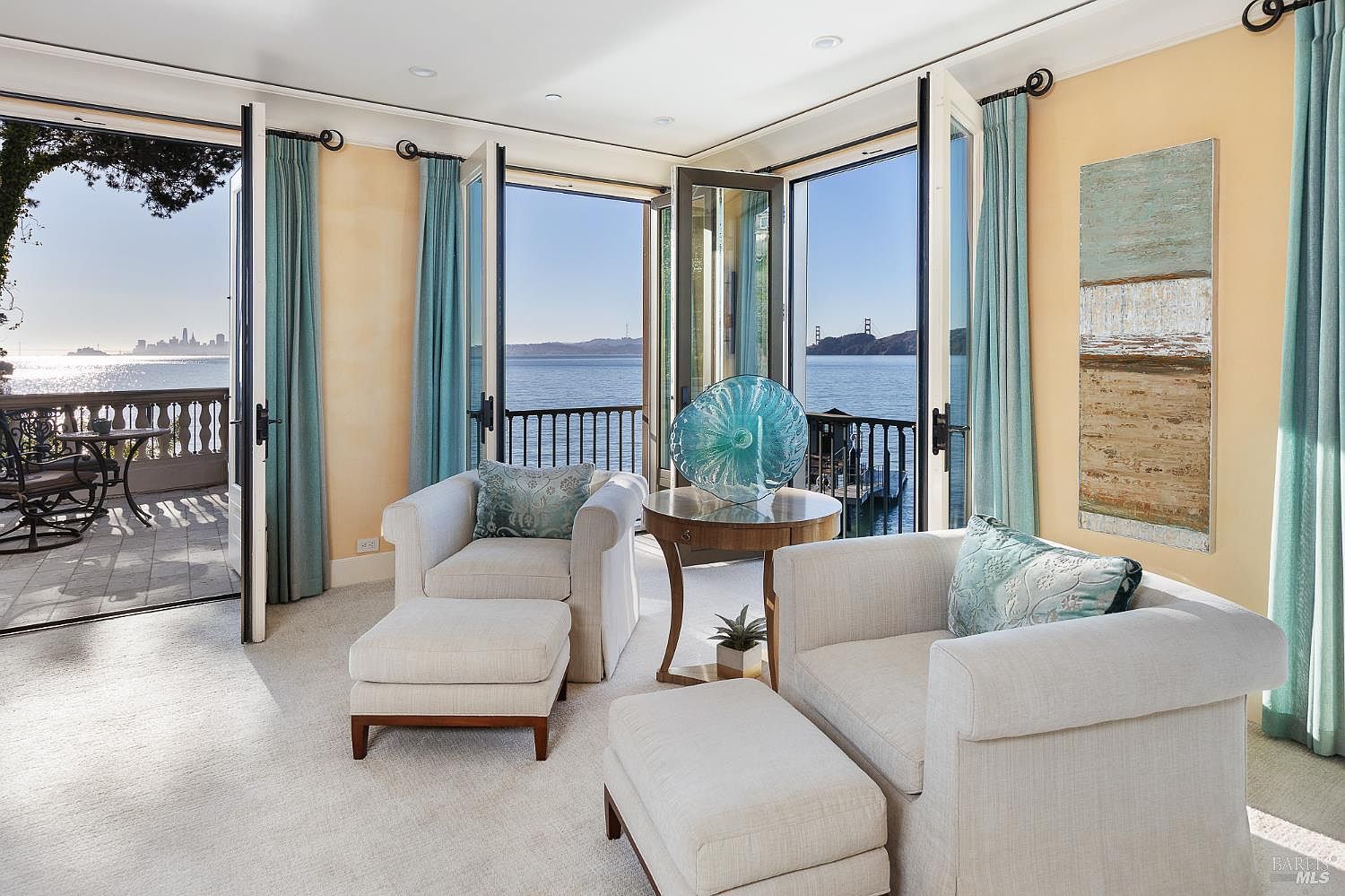 This bright and airy living space features two comfortable armchairs with matching ottomans, centered around a small round table topped with a decorative glass sculpture. Large glass doors open onto a private balcony, framing expansive, panoramic views of the bay, the city skyline, and the Golden Gate Bridge. The room is decorated in a soft, neutral palette with light-toned carpeting and pale yellow walls, creating a serene and sophisticated atmosphere.
