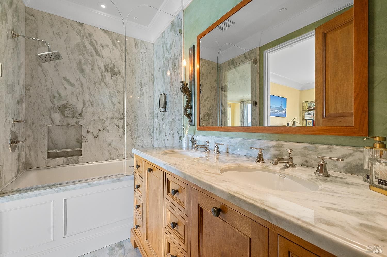 This elegant bathroom features a dual-sink vanity with light wood cabinetry and a light-toned stone countertop. A large, framed mirror hangs above the vanity, reflecting the adjacent room, while a glass-enclosed shower and tub combination is finished with patterned stone wall tiles. The space is accented by green wall paint and classic metal fixtures, creating a sophisticated and bright atmosphere.