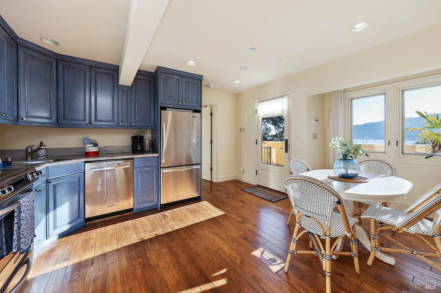 This bright kitchen features deep blue cabinetry paired with stainless steel appliances and a neutral countertop. A round dining table with woven-style chairs sits near large windows that provide a scenic view of the water, while warm-toned wood flooring runs throughout the space. The room is illuminated by recessed lighting and natural light streaming in from the adjacent door and windows, creating an inviting and airy atmosphere.