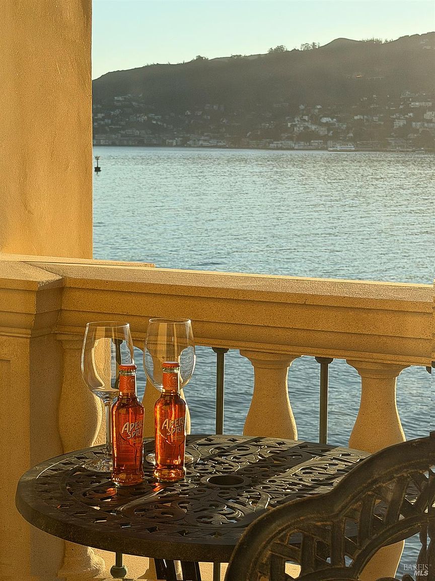 This balcony offers a serene, elevated view of a calm bay with rolling hills in the distance, captured during the golden hour. In the foreground, a decorative wrought-iron bistro table holds two glasses and two bottles of Aperol Spritz, creating an inviting and relaxed atmosphere. The stone balustrade adds a classic architectural touch, framing the picturesque waterfront vista.
