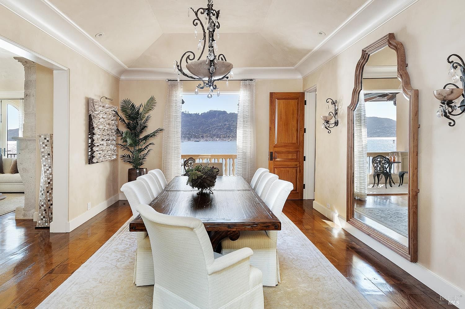 This elegant dining room features a large, dark wood table surrounded by white upholstered chairs, set beneath a decorative chandelier. The space is framed by light-colored walls, a large arched mirror, and a wide opening that reveals a scenic waterfront view through a balcony. The room exudes a sophisticated and airy atmosphere, enhanced by the polished wood flooring and a neutral area rug.