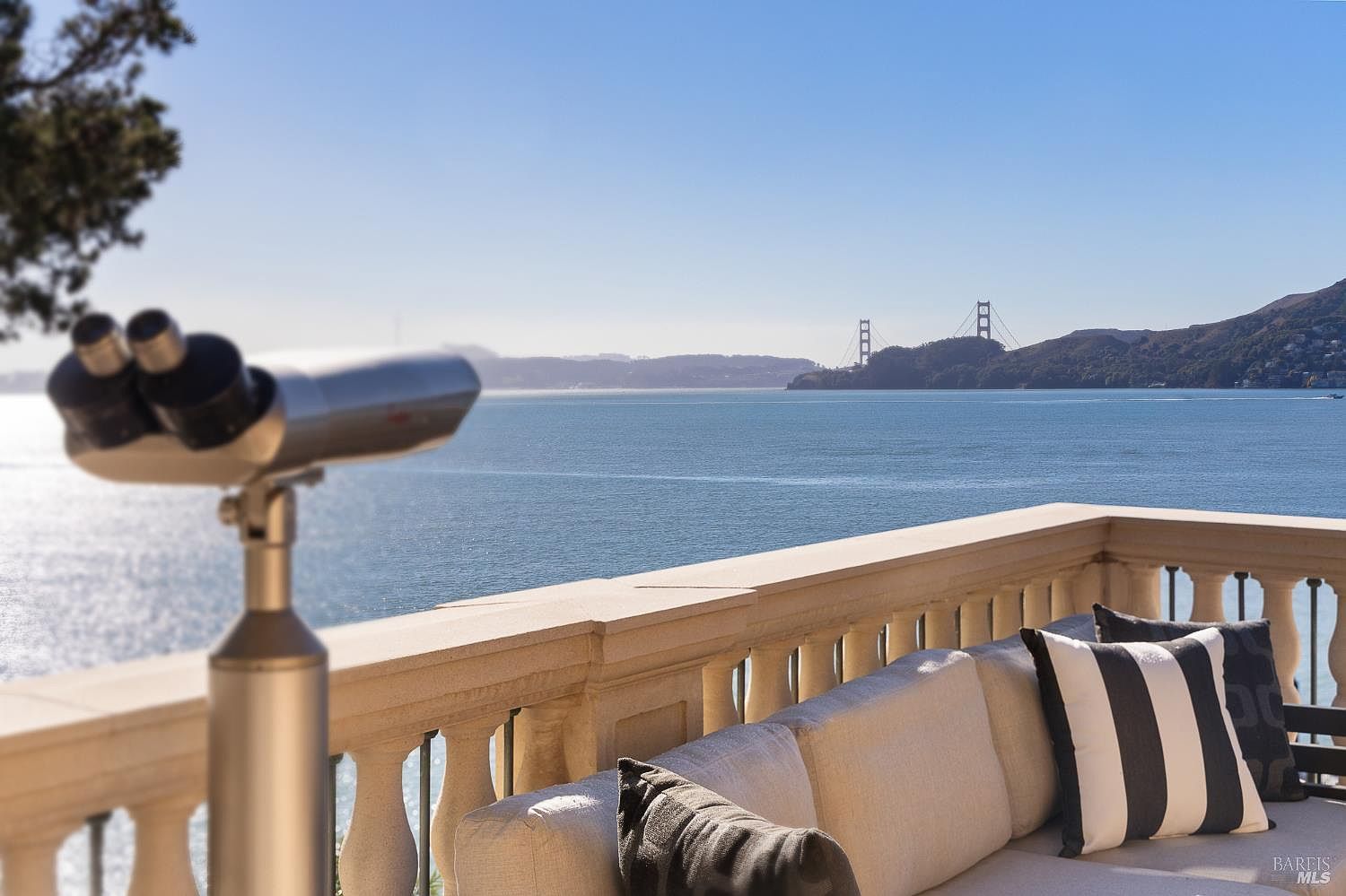 This balcony offers a stunning, elevated view of the bay and the Golden Gate Bridge, framed by a classic stone balustrade. In the foreground, a comfortable outdoor sofa with striped cushions provides a relaxing seating area, while a mounted telescope invites residents to enjoy the scenic vista. The perspective captures the serene atmosphere of a high-end waterfront property, emphasizing the seamless connection between indoor comfort and the expansive natural landscape.
