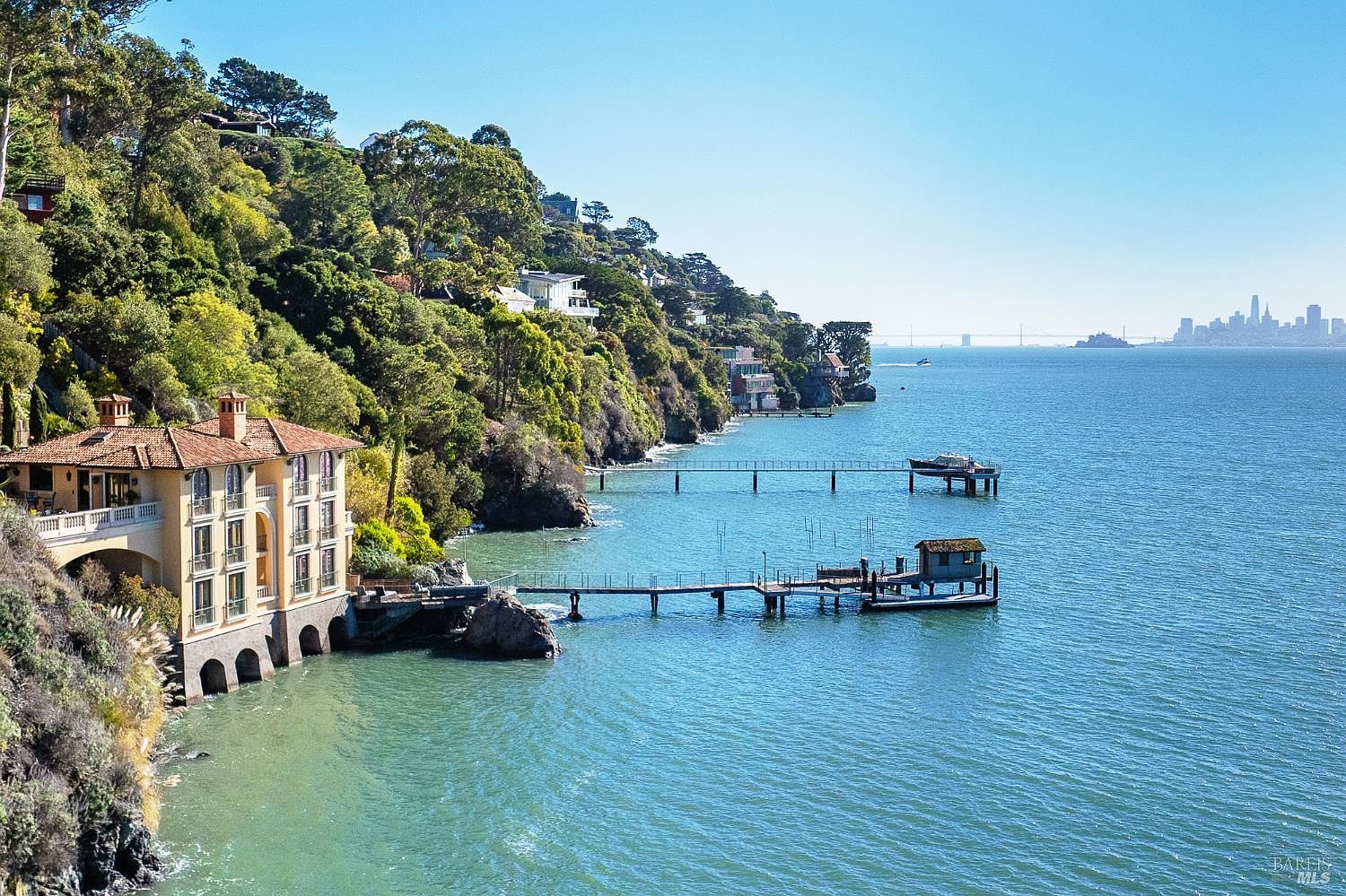 This elevated perspective captures a Mediterranean-style villa perched on a rugged, tree-lined cliff overlooking a vast bay. The property features a multi-level residence with a terracotta-tiled roof, private piers extending into the water, and a scenic backdrop of a distant city skyline. The composition emphasizes the dramatic coastal setting and the seamless integration of luxury architecture with the natural landscape.