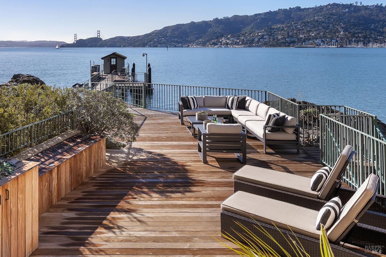 This expansive wooden deck offers a stunning waterfront view, featuring a comfortable outdoor sectional sofa and two lounge chairs positioned to overlook the bay. A private pier with a small structure extends into the water, while the Golden Gate Bridge is visible in the distance against a clear blue sky. The setting provides a serene and luxurious atmosphere, perfect for outdoor relaxation and entertaining.
