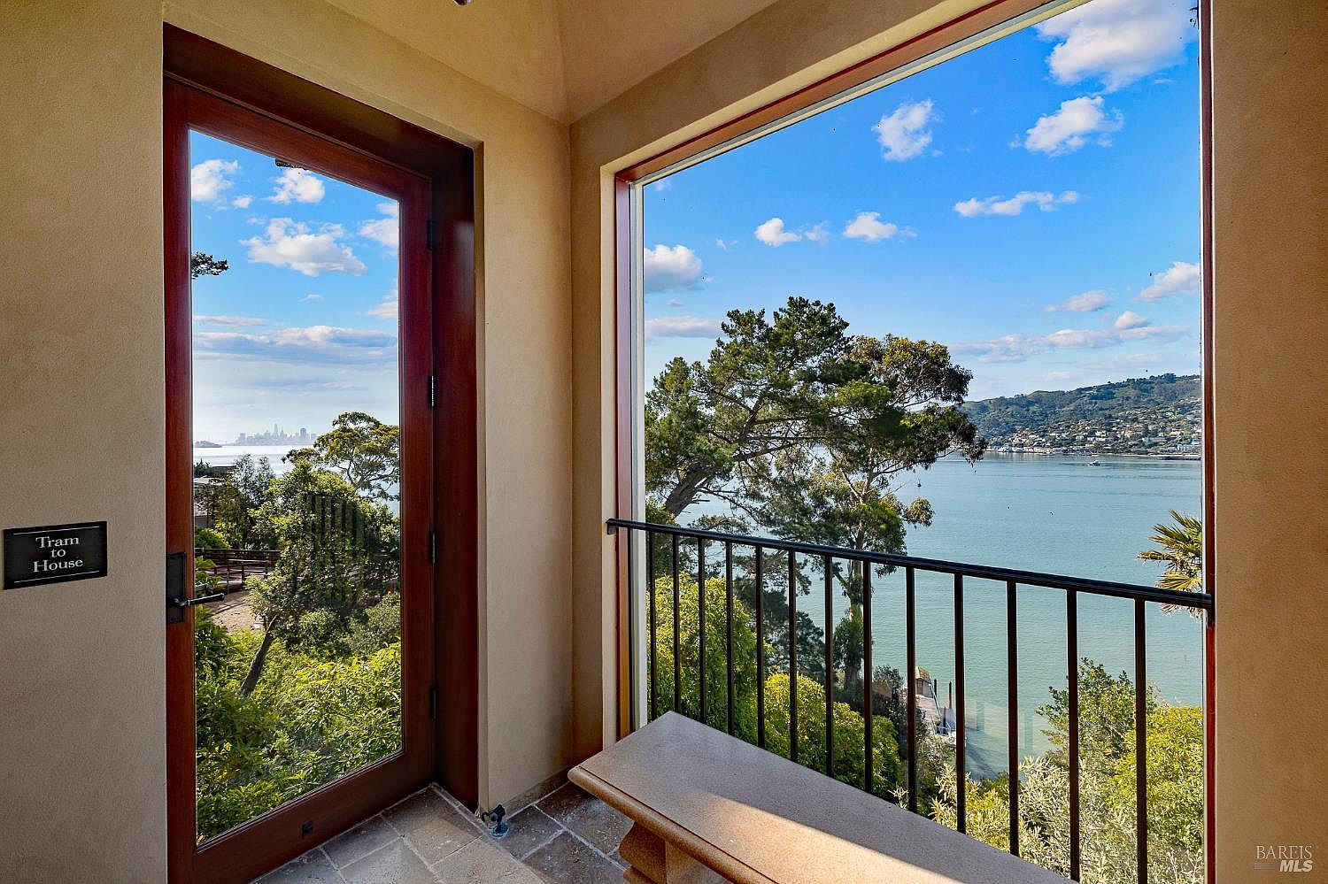 This interior space features a glass door leading to a tram and a large window offering a panoramic view of the bay and distant city skyline. A stone bench sits in the foreground, providing a place to enjoy the natural light and scenic surroundings. The room has a neutral, warm-toned palette that emphasizes the connection between the indoor area and the expansive outdoor landscape.