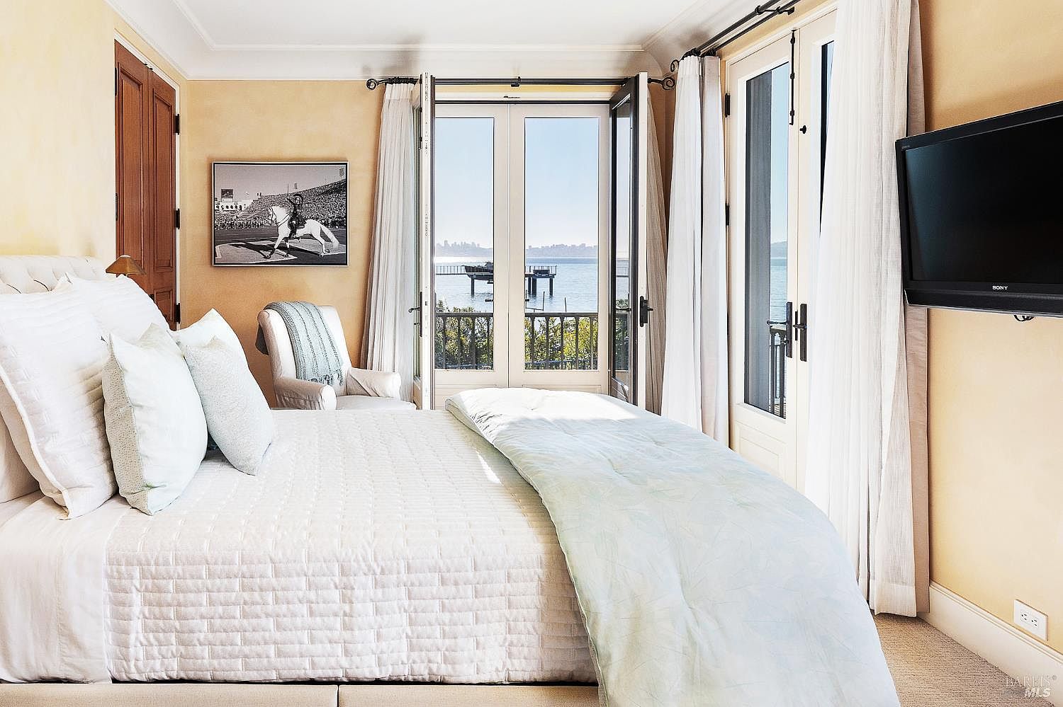This serene primary bedroom features a plush bed with light-colored bedding, positioned to face a set of double doors that open onto a balcony with water views. The room is decorated in a soft, neutral palette with a framed equestrian photograph on the wall and a comfortable armchair in the corner. A wall-mounted television is positioned to the right, and the space is filled with natural light streaming through the glass doors.