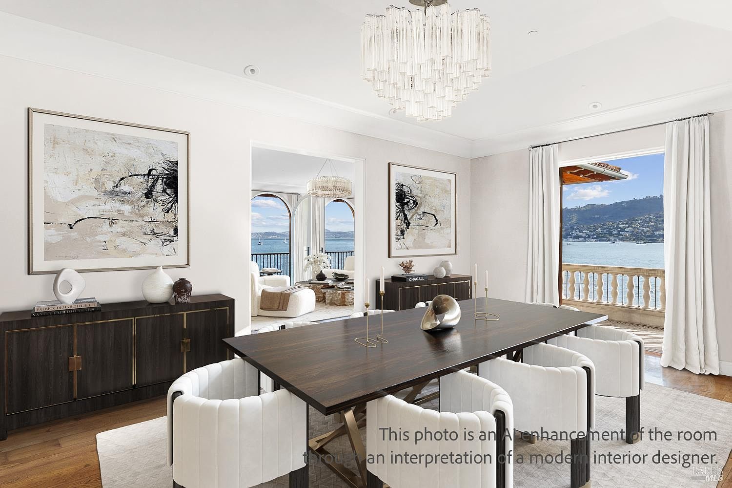 This elegant dining room features a dark wood table surrounded by white upholstered chairs, set beneath a striking crystal chandelier. The space is framed by large windows and a doorway that provide expansive views of the water and distant hills. Two abstract art pieces adorn the white walls, while dark wood sideboards provide sophisticated storage, creating a refined and airy atmosphere.