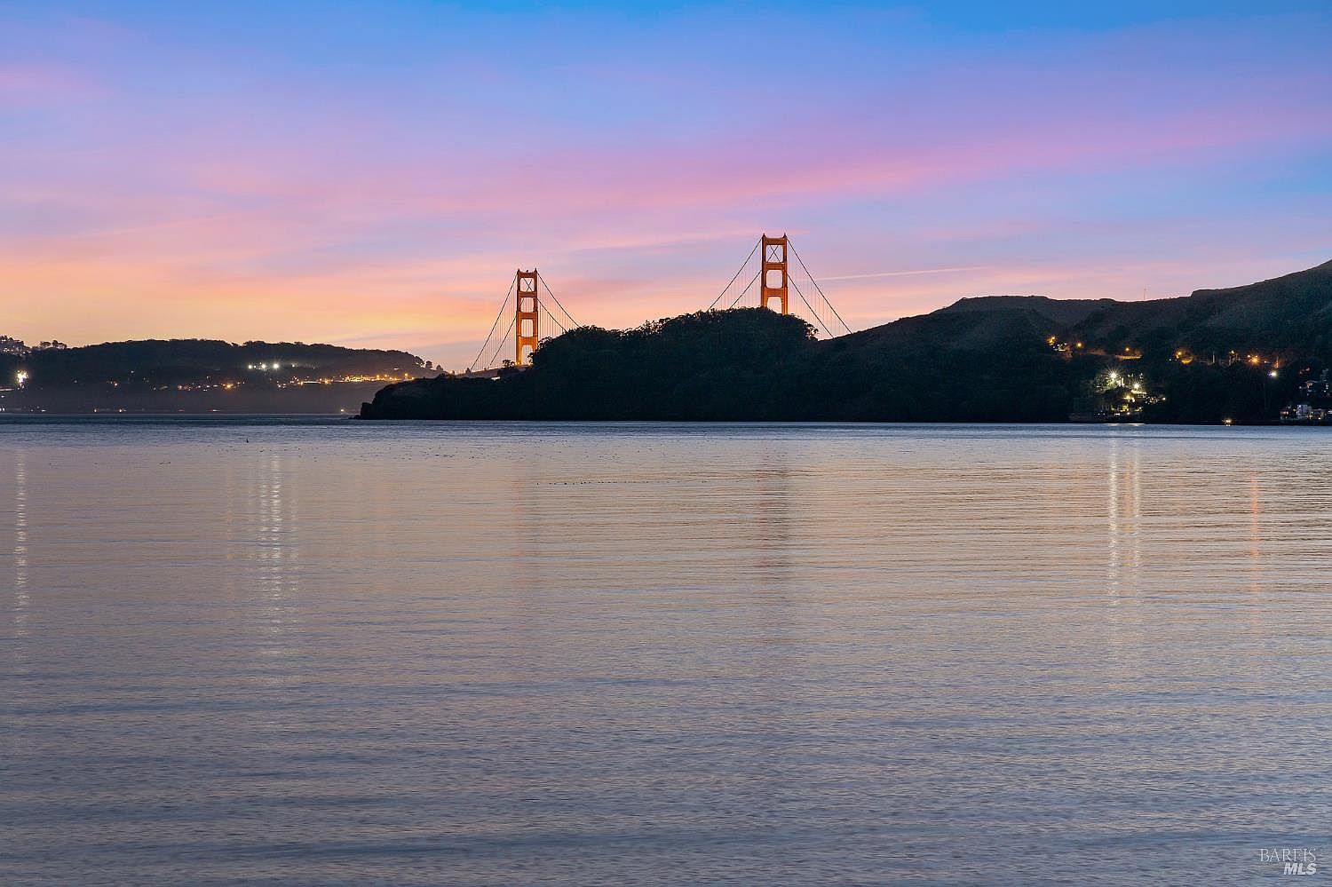 This scenic landscape shot captures the iconic Golden Gate Bridge at dusk, framed by a dark, silhouetted headland in the foreground. The sky is painted with soft hues of pink, purple, and blue, reflecting gently on the calm, expansive waters of the bay. The composition emphasizes the bridge's distinctive towers as a focal point, highlighting the serene and picturesque nature of the surrounding coastal environment.