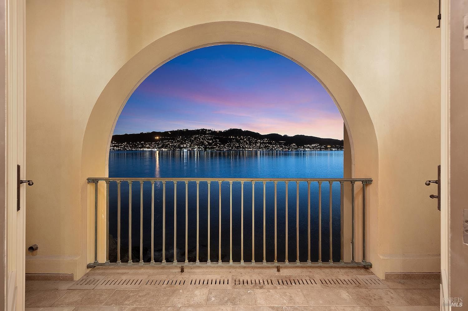 This image captures a stunning, arched balcony opening that frames a panoramic view of a tranquil lake and distant hillside at twilight. The balcony features a classic metal railing and stone flooring, creating a sophisticated transition between the interior space and the outdoor scenery. The composition emphasizes the dramatic architectural archway, which serves as a natural frame for the vibrant sunset colors reflecting off the water.