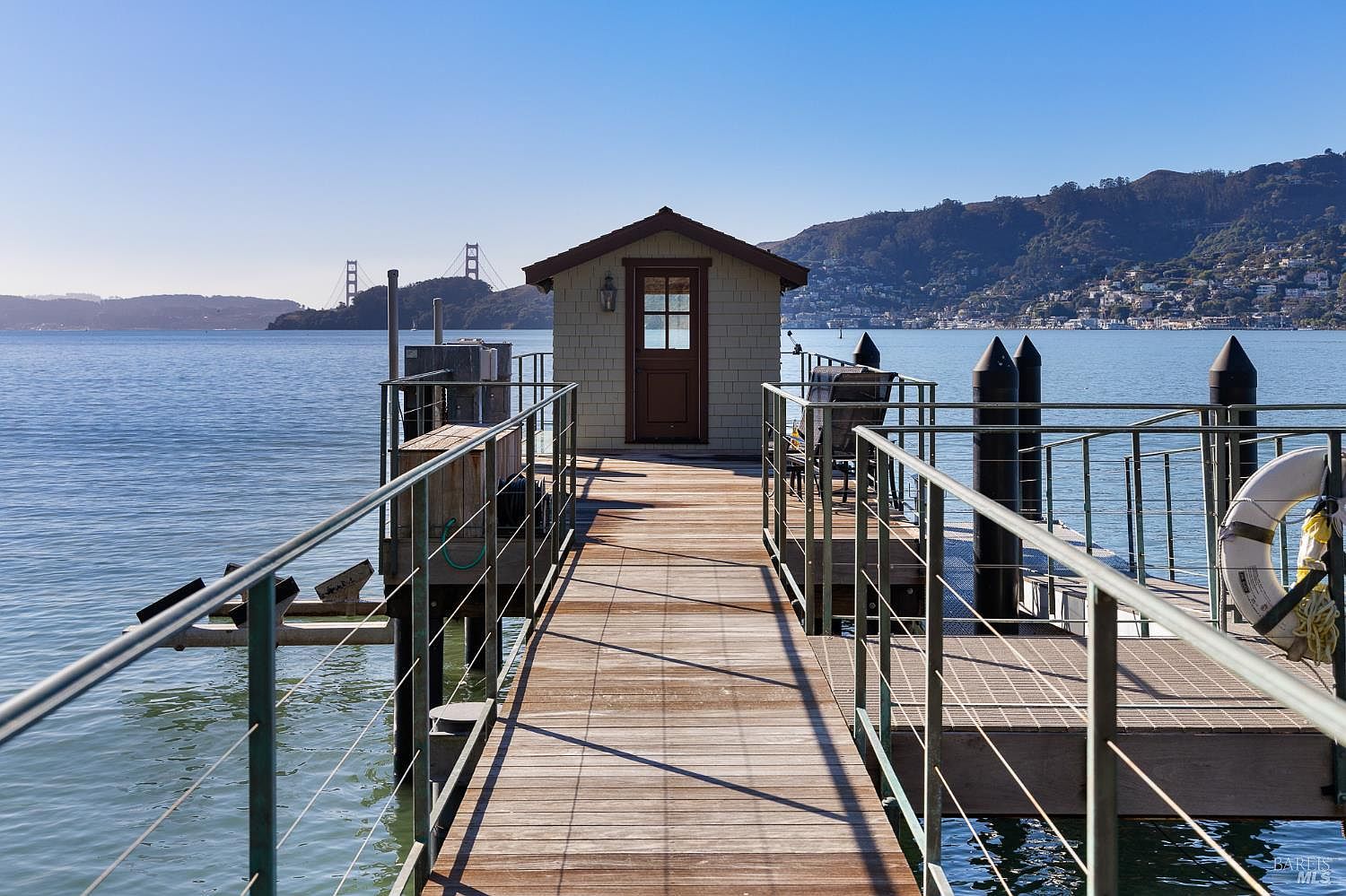 This image captures a wooden pier extending over calm water, leading to a small, shingled structure with a single door. The perspective is centered, drawing the eye toward the building while framing the distant Golden Gate Bridge and hillside homes in the background. Metal railings line the walkway, and a life ring is mounted on the right, emphasizing the nautical setting and serene waterfront lifestyle.