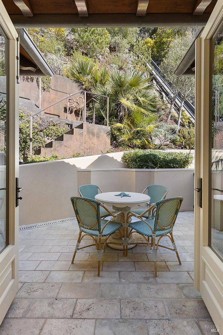 This inviting outdoor patio is framed by open French doors, creating a seamless transition from the interior to the exterior living space. The area features a round stone-topped table surrounded by four woven bistro-style chairs, set upon a light-toned stone paved floor. Beyond the low retaining wall, a tiered hillside garden with lush greenery and a set of stairs provides a serene, private backdrop.