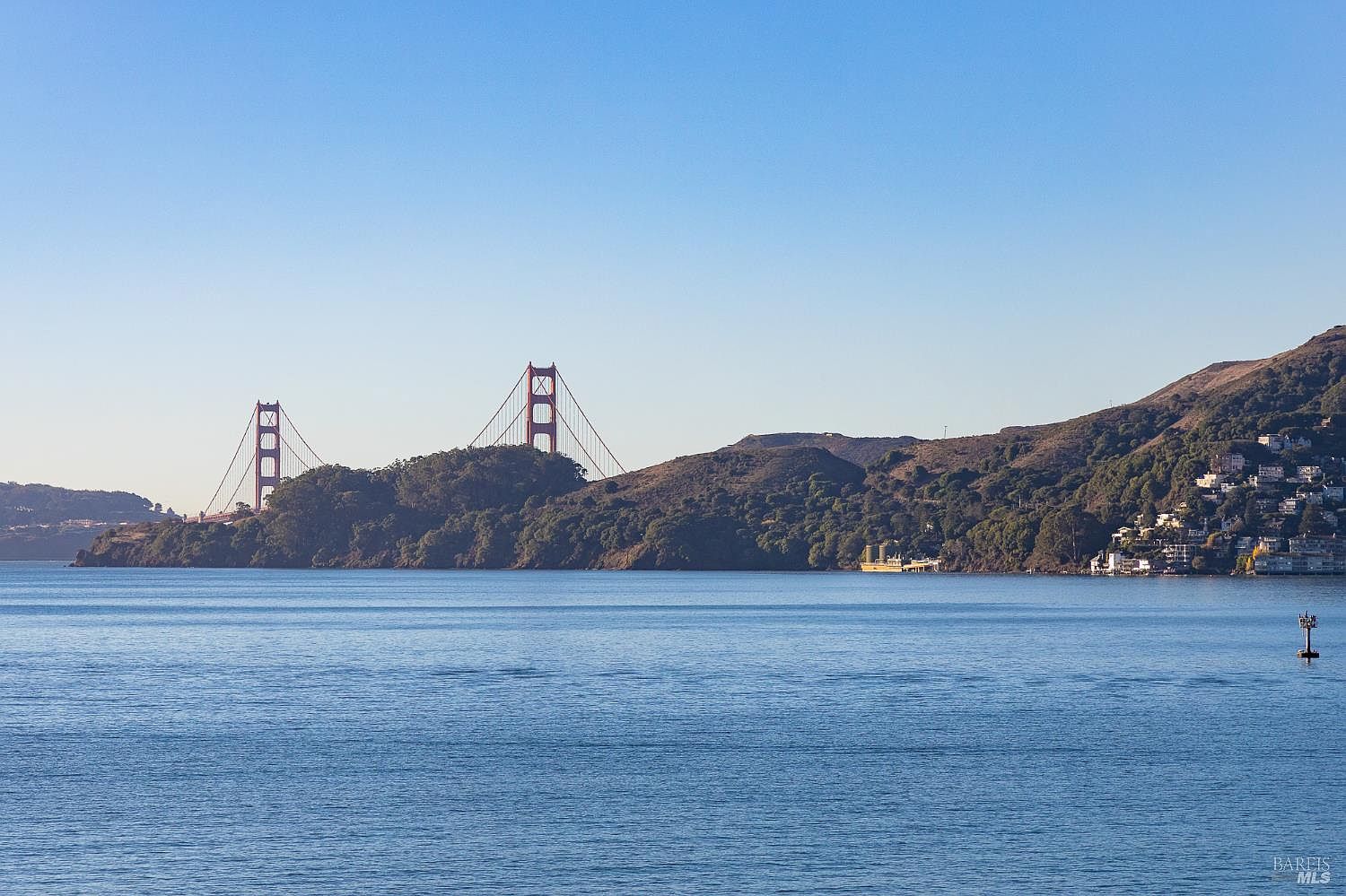 This scenic landscape shot captures a wide-angle view of the iconic Golden Gate Bridge spanning across the water, framed by rolling, tree-covered hills. In the foreground, the calm, deep blue water stretches across the frame, while the right side of the image reveals a hillside residential area with homes nestled among the greenery. The perspective is elevated and expansive, emphasizing the natural beauty and prime location of the coastal setting.
