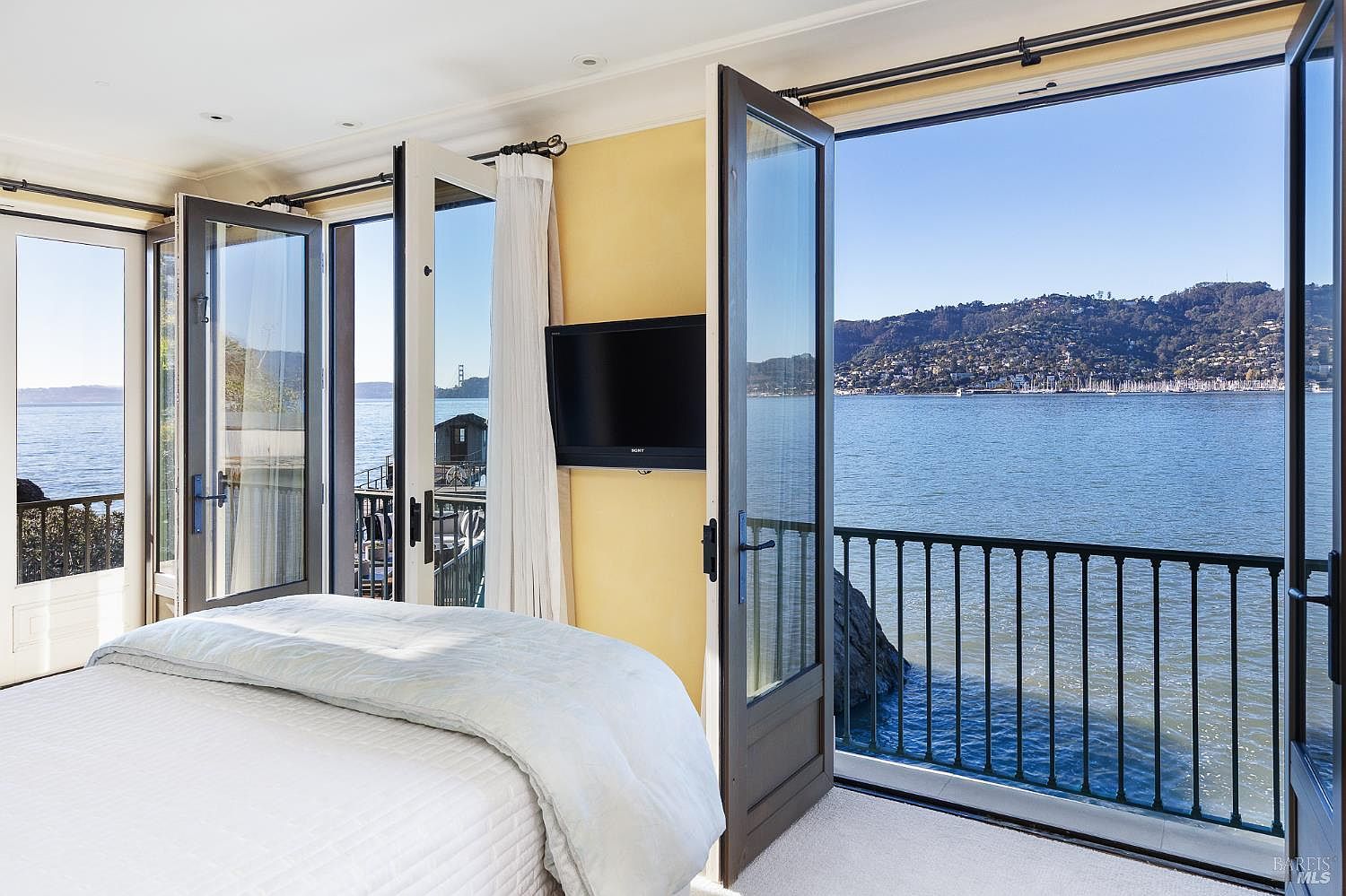 This primary bedroom features a serene, light-filled atmosphere with direct access to a private balcony overlooking a scenic bay. The room is anchored by a comfortable bed with a light-colored quilt, positioned to take full advantage of the expansive water views through large glass doors. A wall-mounted television is centered on a soft yellow wall, creating a cozy and relaxing retreat with a strong connection to the outdoors.