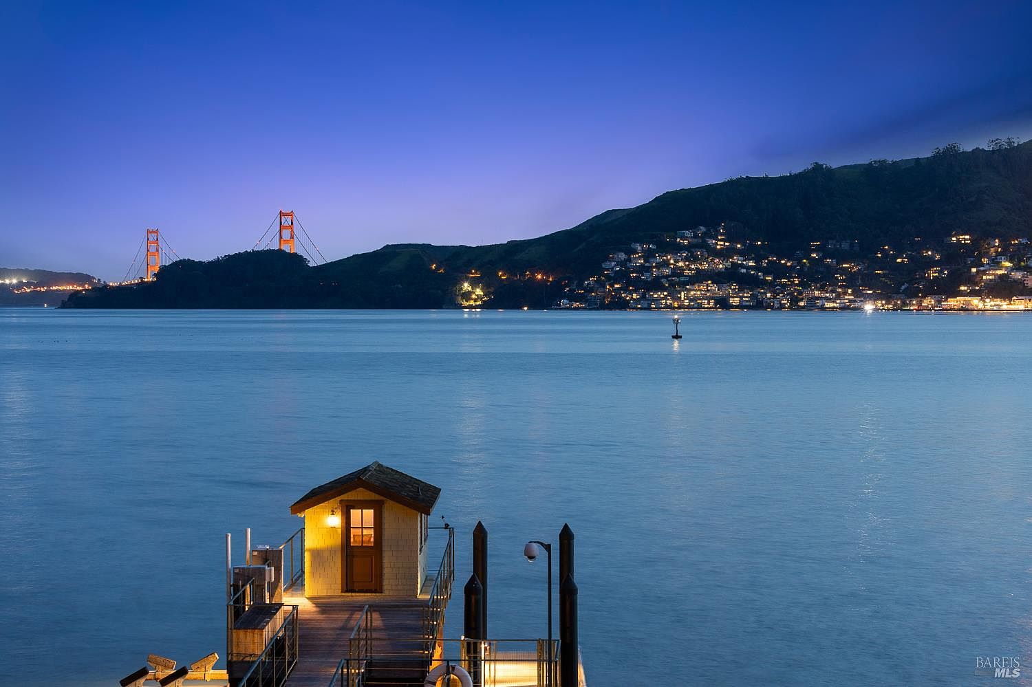 This cinematic shot captures a private wooden pier extending into a calm bay at twilight, featuring a small, illuminated structure at the end of the dock. In the distance, the iconic Golden Gate Bridge is silhouetted against a deep blue sky, while the hillside across the water is dotted with the warm, twinkling lights of a residential community. The perspective is elevated, looking down the length of the pier toward the expansive water and the scenic coastal landscape beyond.