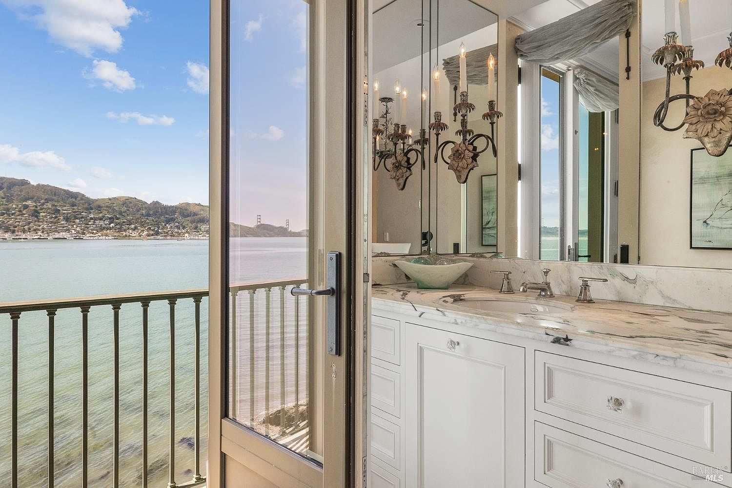 This elegant bathroom features a white vanity with a stone countertop and a unique glass vessel sink, complemented by a large mirror that reflects the room's sophisticated lighting. A glass door opens directly onto a balcony, providing a stunning, unobstructed view of the bay and distant hills. The combination of classic cabinetry and the dramatic waterfront vista creates a luxurious and serene atmosphere.