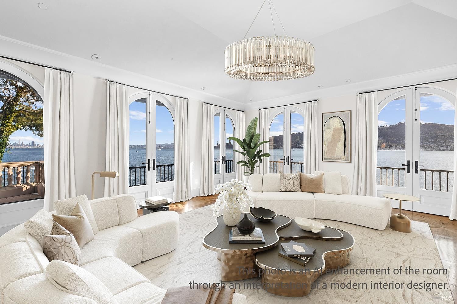 This bright and airy living room features a sophisticated, neutral color palette with two curved, cream-colored sofas arranged around a unique, multi-tiered organic-shaped coffee table. Large arched windows and glass doors line the walls, providing expansive, unobstructed views of the bay and city skyline. A grand, circular crystal chandelier serves as a focal point, while the minimalist decor and light-toned area rug create an atmosphere of refined luxury.