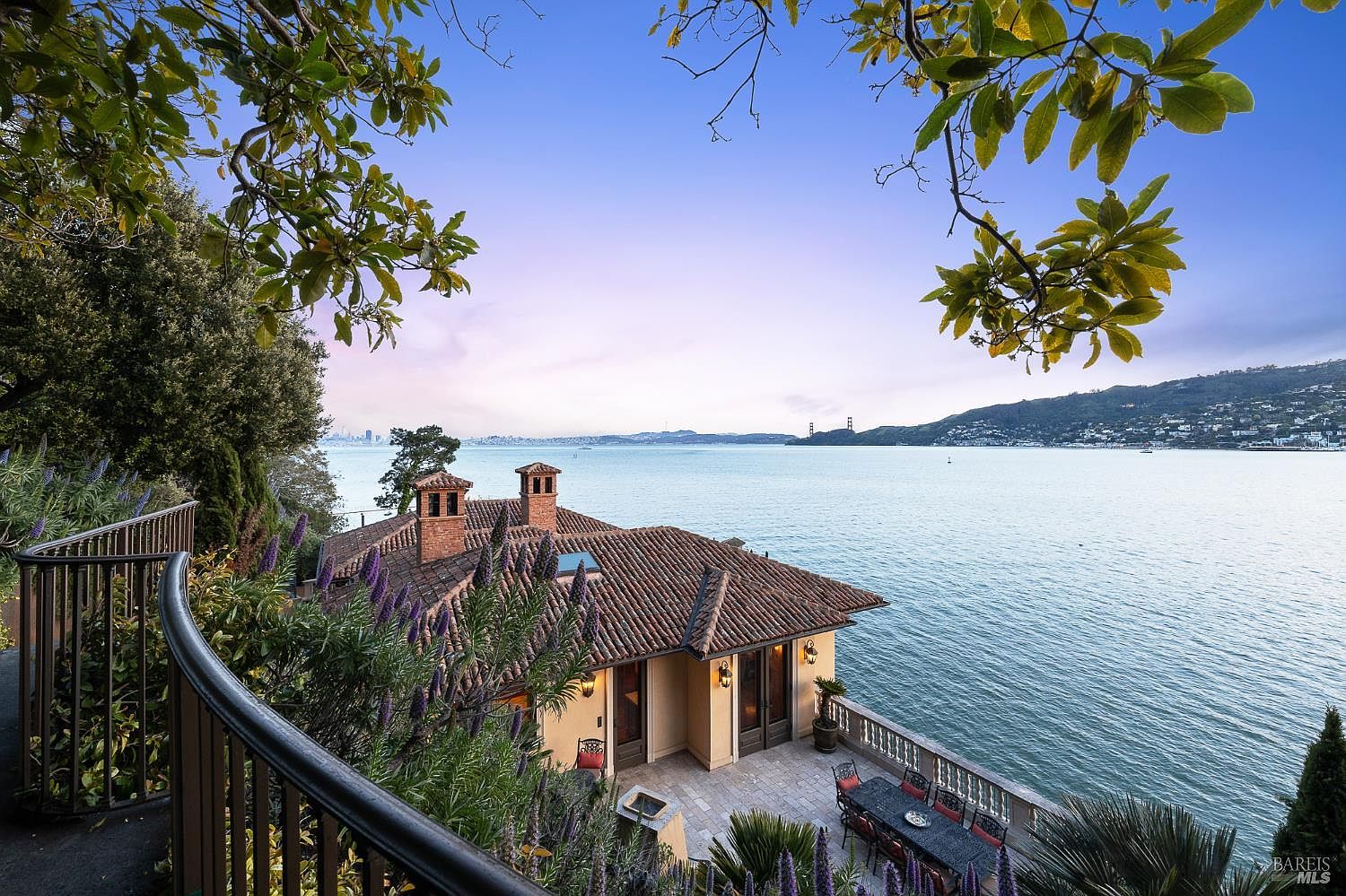 This elevated perspective captures a stunning Mediterranean-style home situated directly on the waterfront, featuring a classic terracotta tile roof and warm-toned stucco walls. A spacious stone-paved patio below offers an ideal outdoor dining area, while the foreground includes a curved balcony railing and vibrant purple flowers. The scene is framed by lush tree branches, providing a picturesque view of the bay, distant city skyline, and the iconic Golden Gate Bridge.
