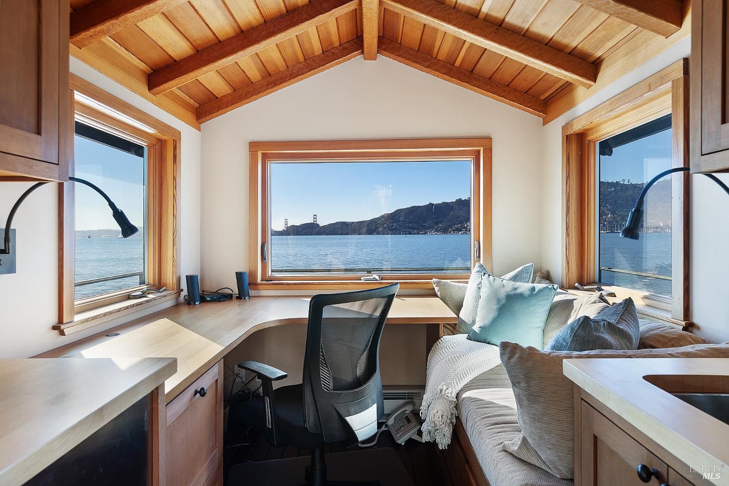 This cozy home office features a built-in corner desk with light wood surfaces, positioned to take full advantage of the panoramic water views through large windows. A comfortable bench seat with plush cushions runs along the right wall, creating a relaxing nook beneath the vaulted, wood-paneled ceiling. The space is bright and airy, offering a serene environment that frames the distant bridge and hillside landscape.