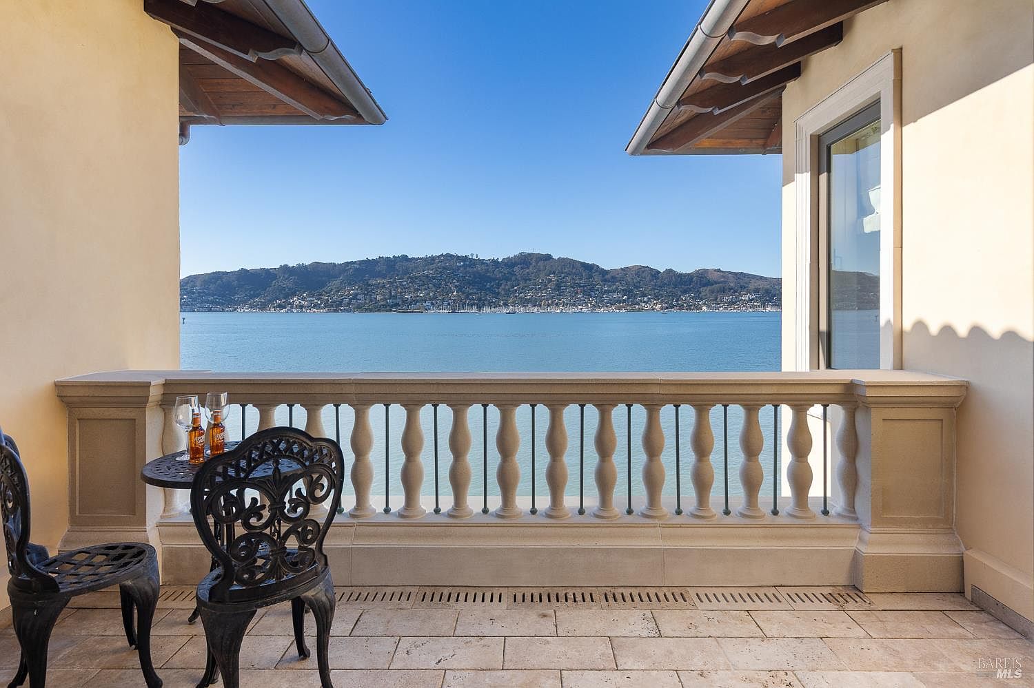 This elegant balcony offers a stunning, unobstructed view of a calm bay and distant hills under a clear blue sky. The space features a classic stone balustrade, light-toned stone flooring, and a small bistro set with two ornate metal chairs. The perspective is framed by the architectural overhangs of the building, creating a serene and sophisticated outdoor retreat.
