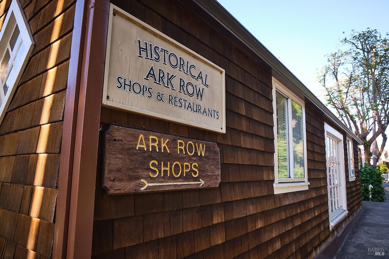 This side view of a historic building features dark brown cedar shake siding and classic white-trimmed windows. Two prominent signs are mounted on the wall, identifying the location as 'Historical Ark Row Shops & Restaurants' and providing directional guidance. The perspective captures the textured facade and the charming, rustic character of the structure under a clear blue sky.