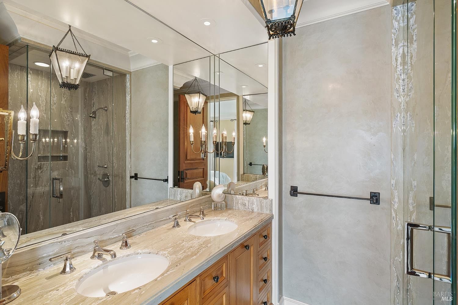This primary bathroom features a double vanity with a light-toned stone countertop and integrated sinks, complemented by warm-toned wood cabinetry. A large mirror spans the wall above the vanity, reflecting the shower area which is enclosed by glass panels and finished with stone-tiled walls. The space is illuminated by elegant wall-mounted light fixtures and recessed ceiling lights, creating a sophisticated and polished atmosphere.