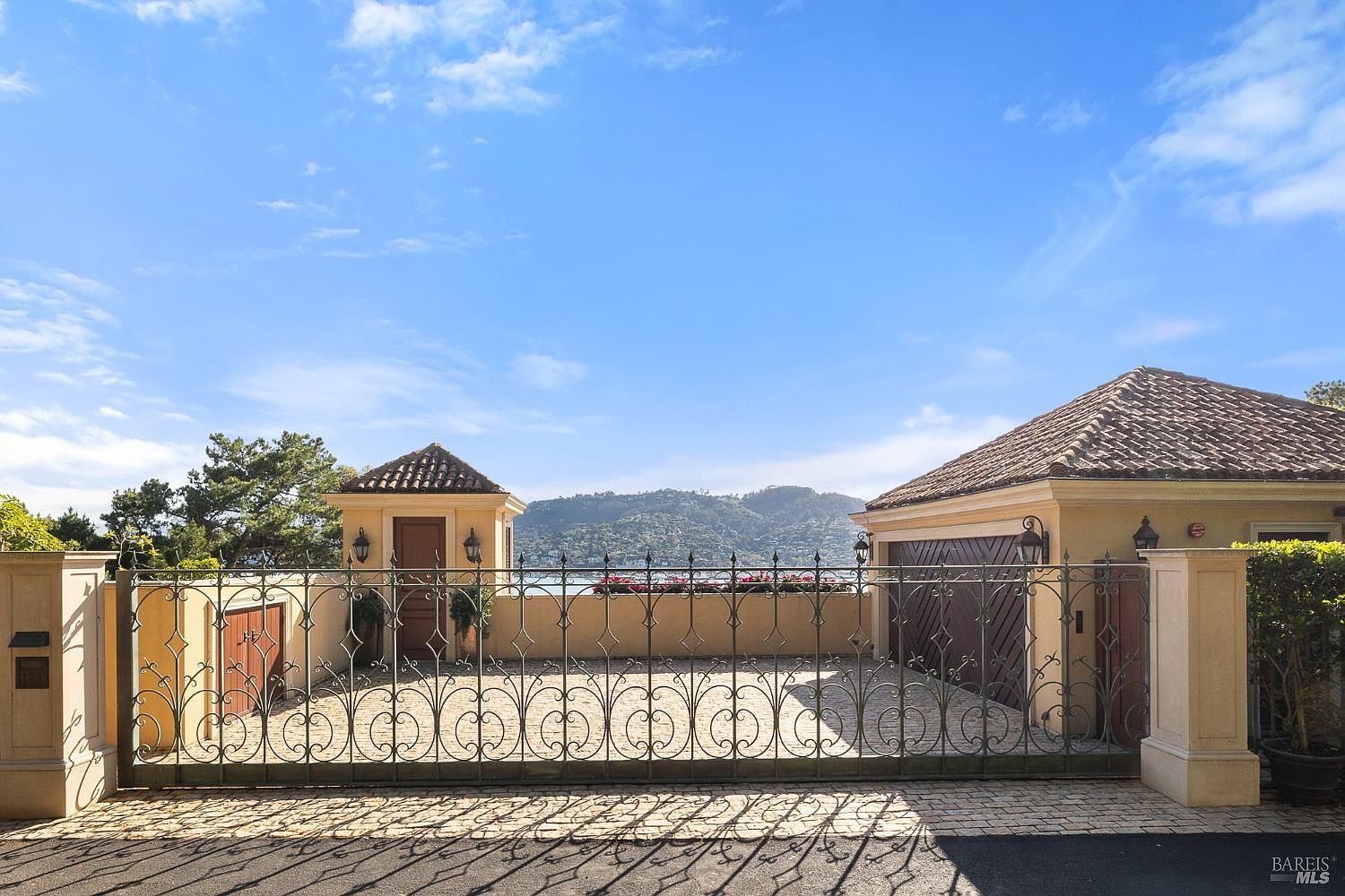 This front-facing view showcases a gated entrance to a Mediterranean-style property, featuring a decorative wrought-iron fence and a paved driveway. The structure includes a small gatehouse on the left and a garage on the right, both topped with terracotta-tiled roofs. Beyond the gate, a scenic view of a body of water and distant hills creates a serene and private atmosphere.