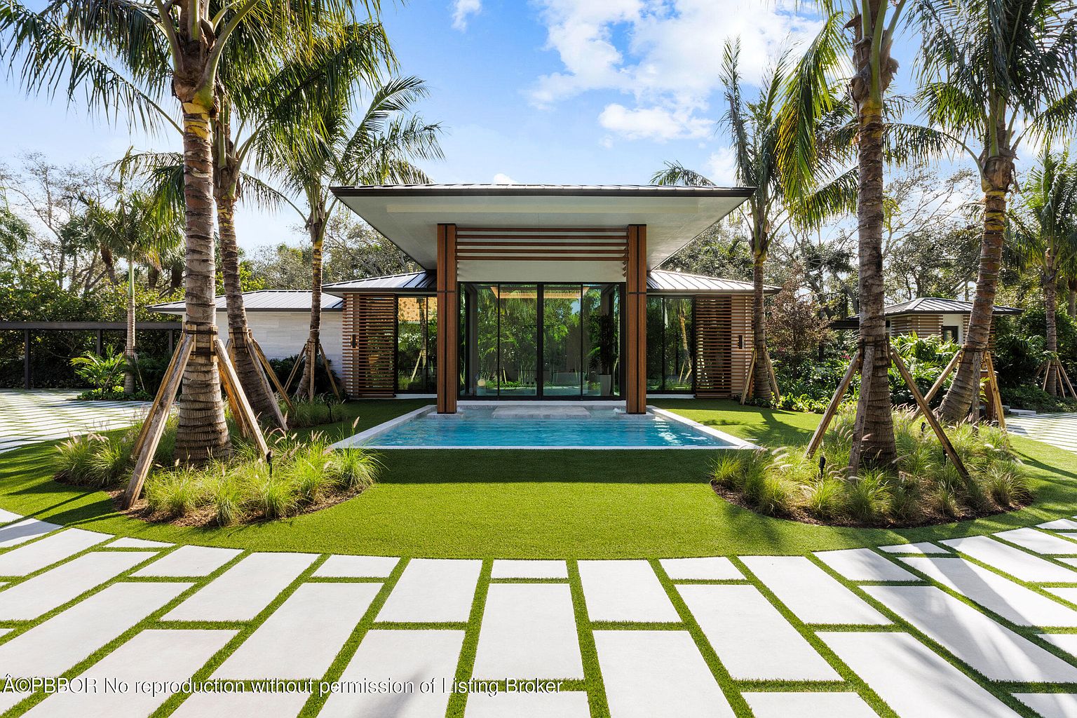 This image showcases a modern home's exterior with a striking pool and spa area. The pool is surrounded by lush green grass and palm trees, creating a tropical oasis. A unique walkway made of large rectangular stones leads to the pool area, enhancing the property's curb appeal and highlighting its luxurious outdoor living space.