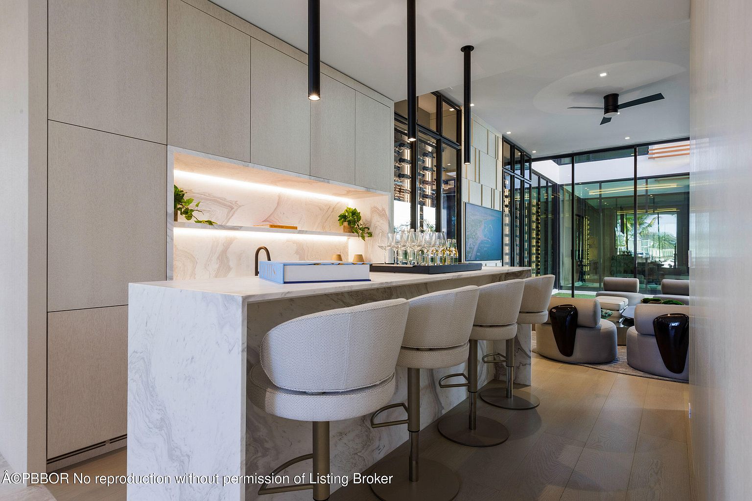 This interior shot showcases a modern wine cellar and bar area, featuring a marble countertop with bar stools, built-in cabinetry with illuminated shelving, and a glass-enclosed wine storage area. The space transitions seamlessly into a living area with comfortable seating, creating an inviting and luxurious atmosphere perfect for entertaining.