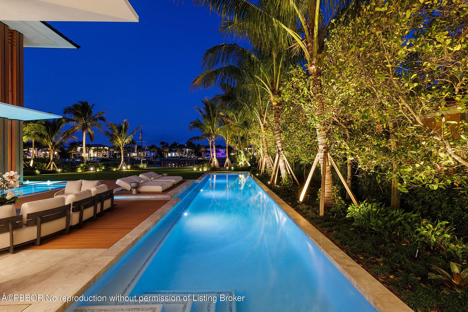 This image showcases a luxurious outdoor pool area at twilight, featuring a long, rectangular pool with illuminated water. Palm trees and manicured greenery line the pool's edge, creating a serene and private atmosphere. Comfortable lounge seating is arranged on a wooden deck adjacent to the pool, suggesting a perfect space for relaxation and entertainment.