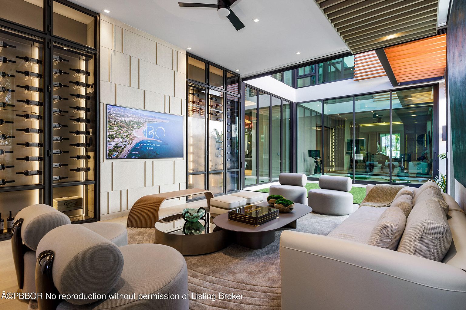 This is a luxurious living room featuring a modern design with a neutral color palette. The room includes a large television, a wine cellar, and comfortable seating arrangements. Large glass windows provide ample natural light and views of the outdoor space, creating an open and airy atmosphere.