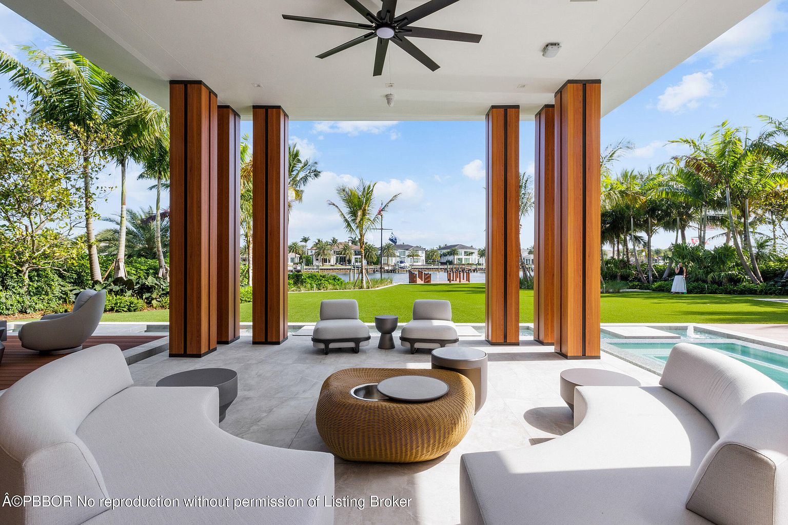 This image showcases a luxurious outdoor living space with a covered patio featuring elegant seating arrangements, including curved sofas and stylish coffee tables. The patio is framed by wooden pillars, offering a view of a well-manicured lawn, palm trees, and waterfront properties in the distance. A pool is visible on the right, adding to the upscale ambiance of this outdoor retreat.