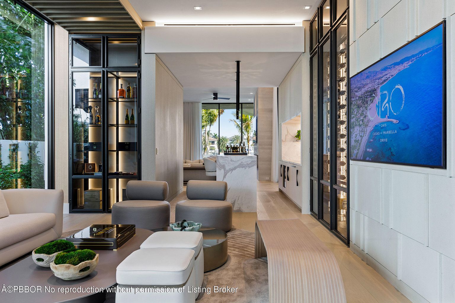 This interior shot showcases a modern living room with a sophisticated design. The room features a sleek display cabinet, comfortable seating arrangements, and a large television displaying an ocean view. The open floor plan leads to a bar area, enhancing the sense of spaciousness and luxury.