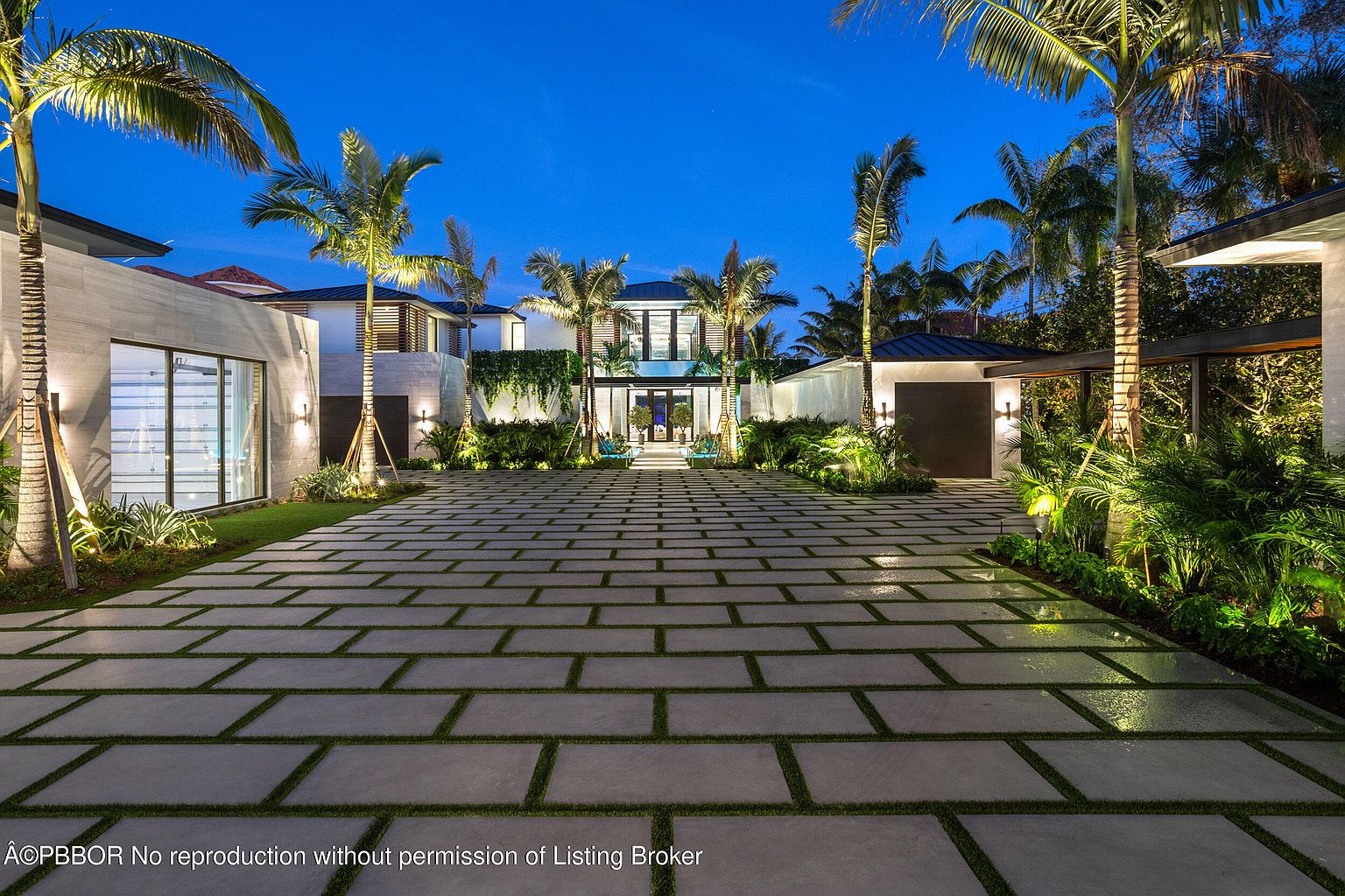 This is a front exterior view of a modern luxury home at dusk, showcasing a large paved driveway with grass accents, leading to a well-lit two-story residence. The property is adorned with palm trees and manicured landscaping, enhancing its curb appeal. The architectural style is contemporary, with clean lines and a focus on outdoor living.