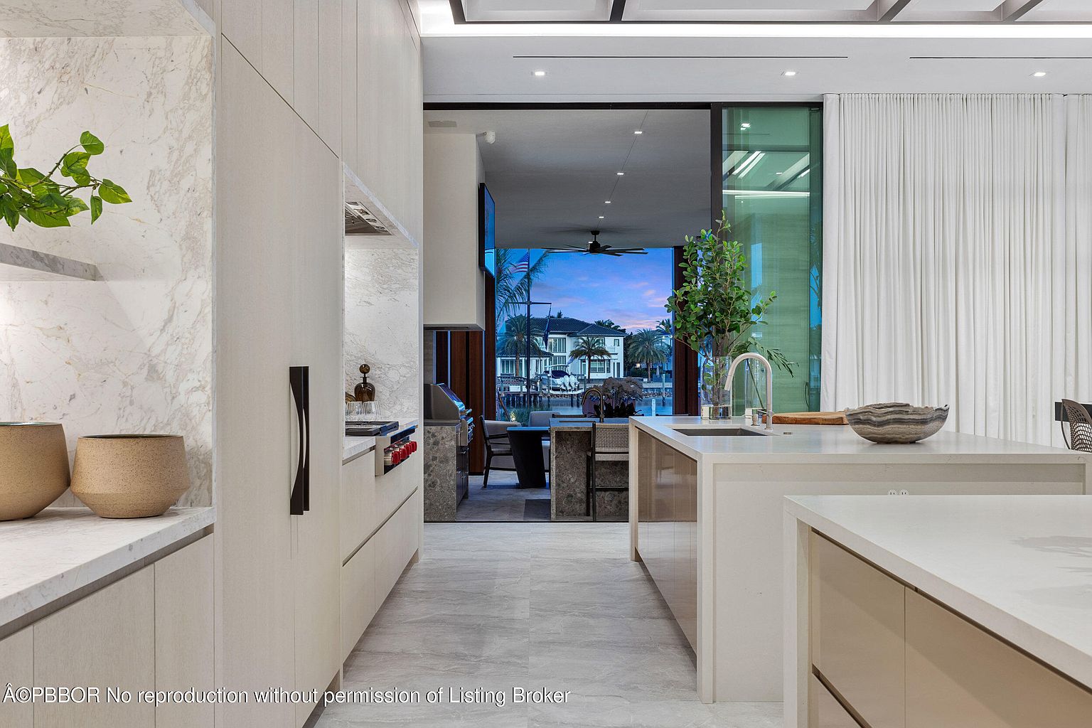 This is a bright, modern kitchen with sleek, light-colored cabinetry and marble countertops. The kitchen features an open layout with a view to an outdoor dining area and waterfront. The design emphasizes clean lines and a minimalist aesthetic, creating a sophisticated and inviting space.
