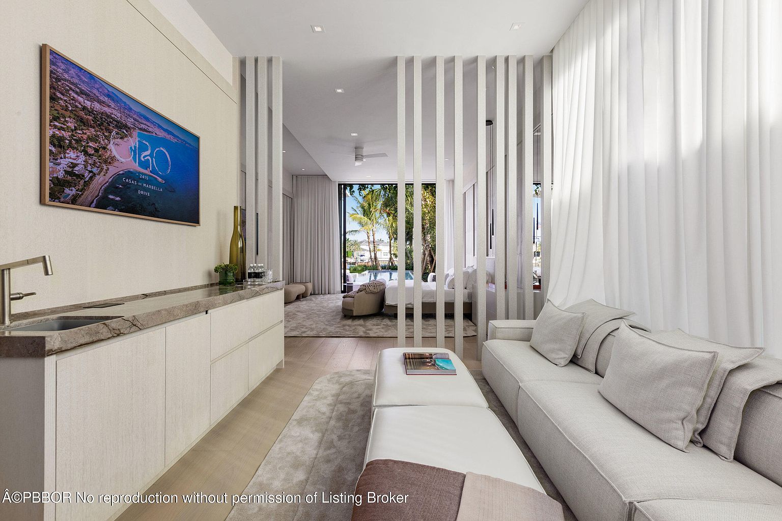 This interior shot showcases a modern living room with a sleek design. A light-colored sofa and ottoman are positioned on a soft rug, complemented by a built-in bar area with a sink and countertop. Vertical slat dividers partially separate the living area from a bedroom space, creating an open yet defined layout, with a TV mounted on the wall.