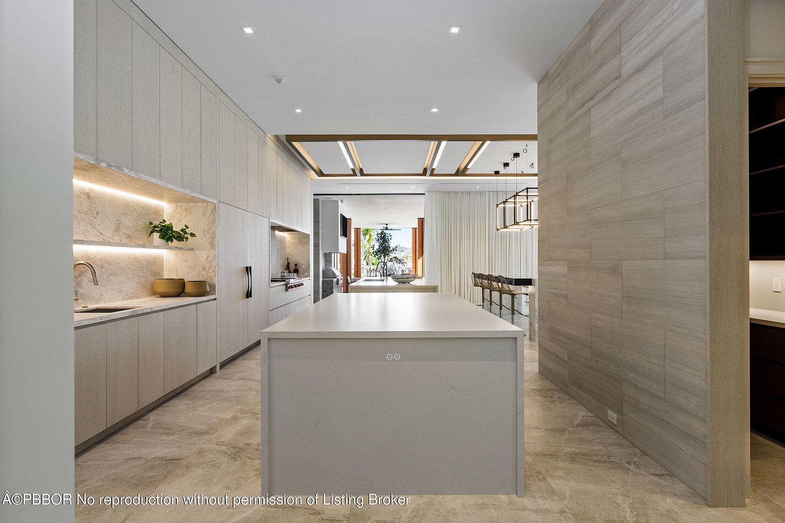 This is a bright, modern kitchen featuring light wood cabinetry and a large central island with a light countertop. The kitchen is open to the dining area, with a view to the outside, and the flooring is a light-colored tile. The overall impression is clean, spacious, and luxurious.