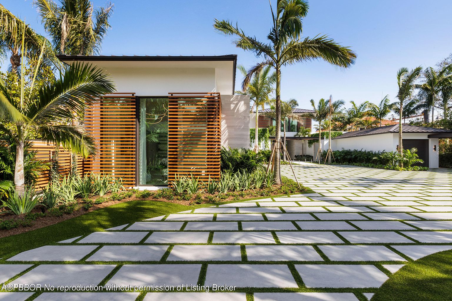 This image showcases the front exterior of a modern home with a striking design. The property features a unique driveway composed of large concrete pavers separated by grass, leading to a contemporary structure with wooden slat accents and lush landscaping. Palm trees and other greenery surround the house, creating a tropical and inviting atmosphere.