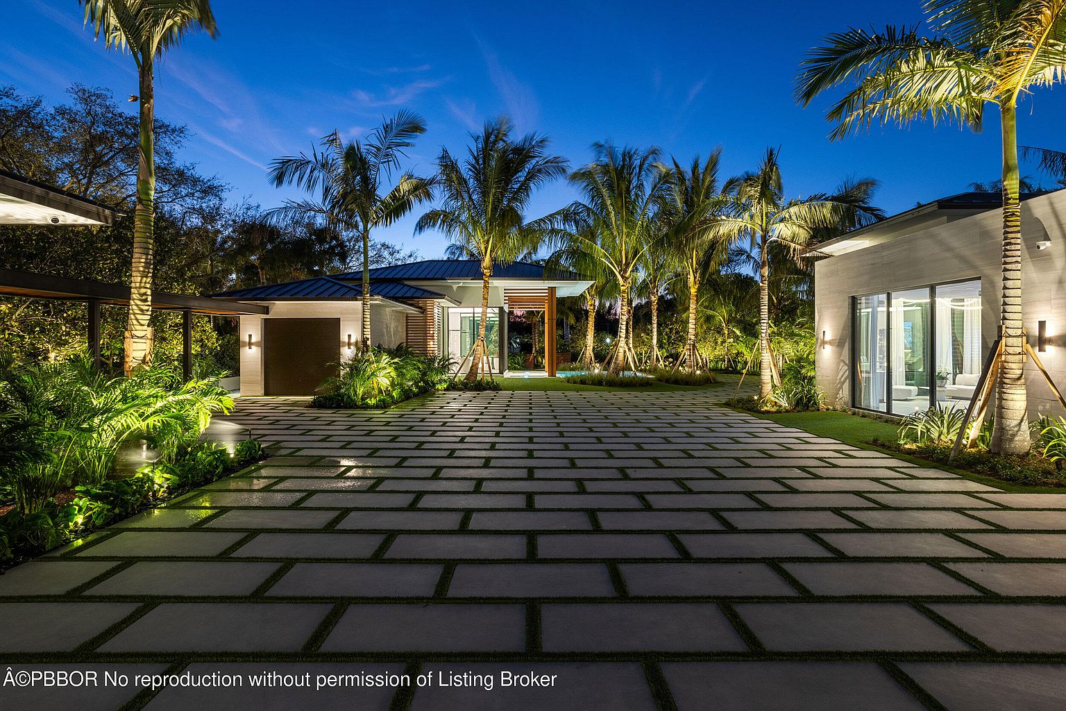 This is a front exterior view of a modern home at dusk, showcasing a wide driveway paved with large rectangular stones and grass in between. The property features lush landscaping with palm trees and illuminated plants, highlighting the architectural details of the house, including clean lines, large windows, and a covered carport. The overall impression is luxurious and inviting.