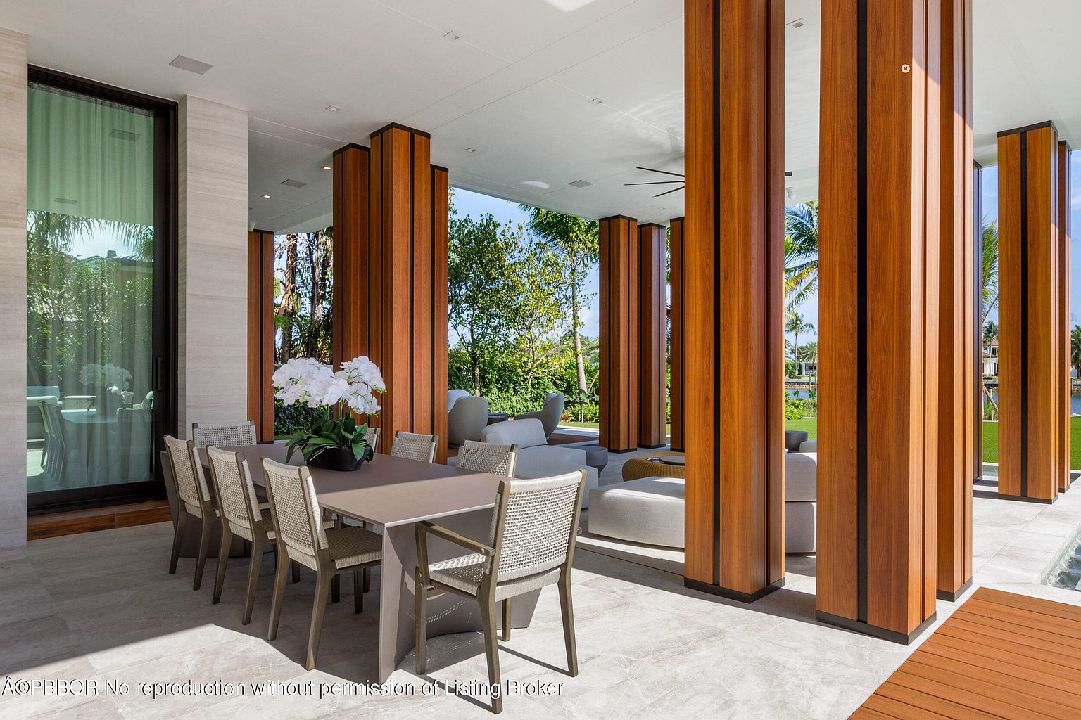This image showcases a luxurious outdoor patio area, featuring a dining table set for eight with a floral centerpiece. The space is defined by striking wooden columns with black accents, providing both structure and visual interest. The patio extends into a comfortable lounge area with plush seating, all set against a backdrop of lush greenery and a glimpse of water, creating an inviting and sophisticated outdoor living space.