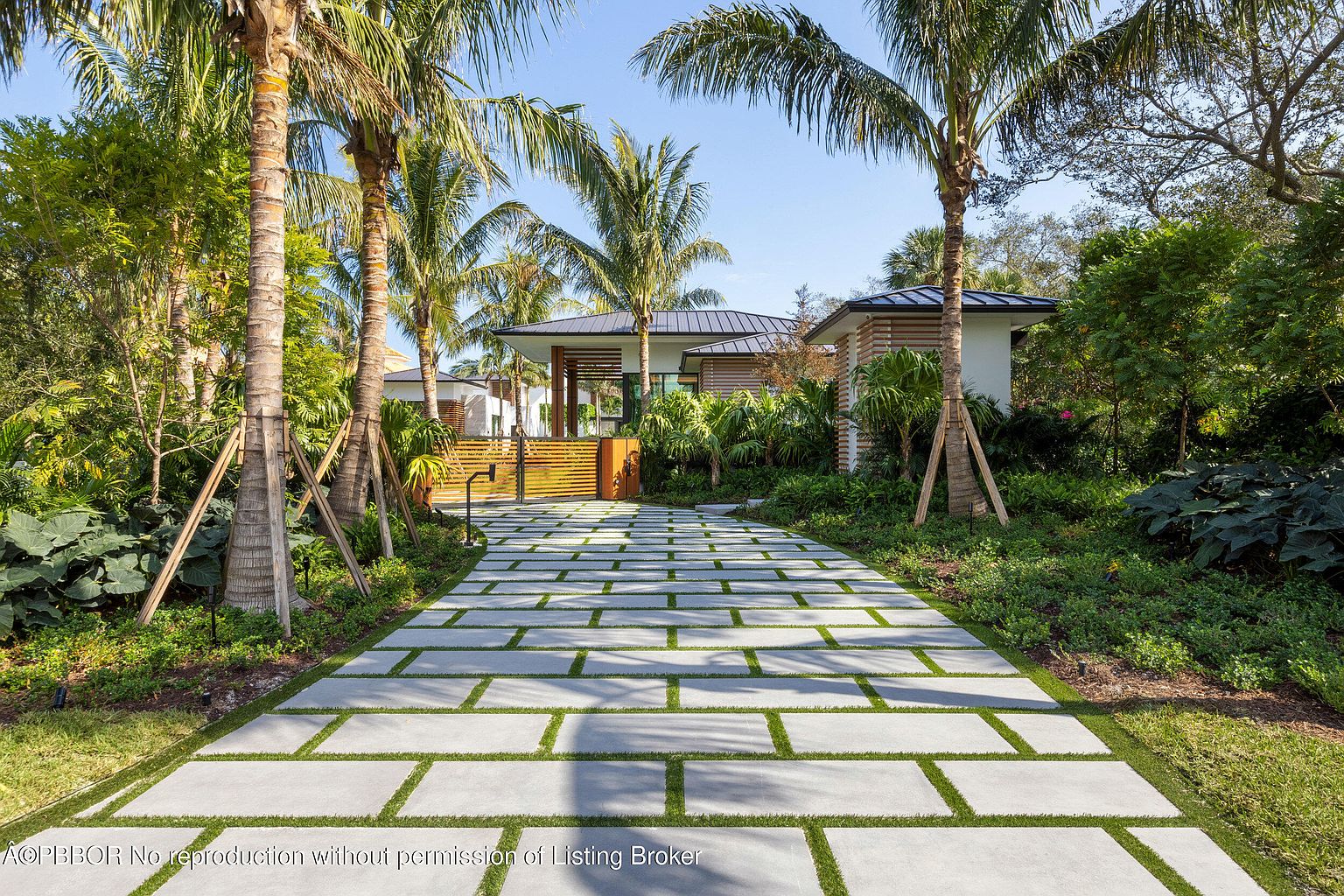 This is a front view of a modern home with a long, paved driveway leading to a gated entrance. The driveway is composed of large, rectangular pavers with grass growing in between, creating a visually appealing pattern. Palm trees line the driveway, adding a tropical feel to the property.