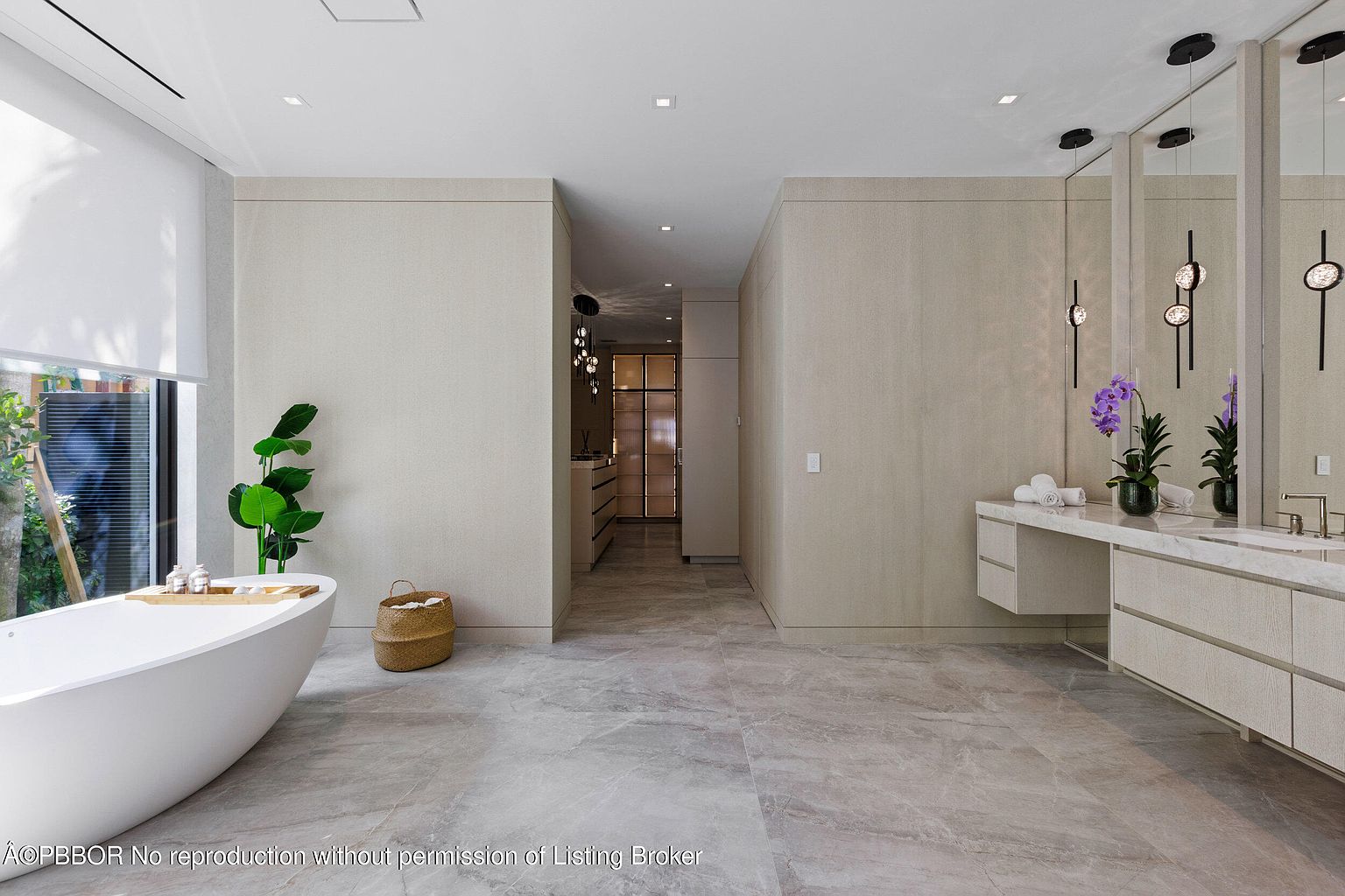 This is a luxurious primary bathroom featuring a freestanding white bathtub positioned near a large window with natural light. The bathroom has a double vanity with modern fixtures, sleek mirrors, and elegant lighting. The neutral color palette and marble flooring create a serene and spa-like atmosphere.