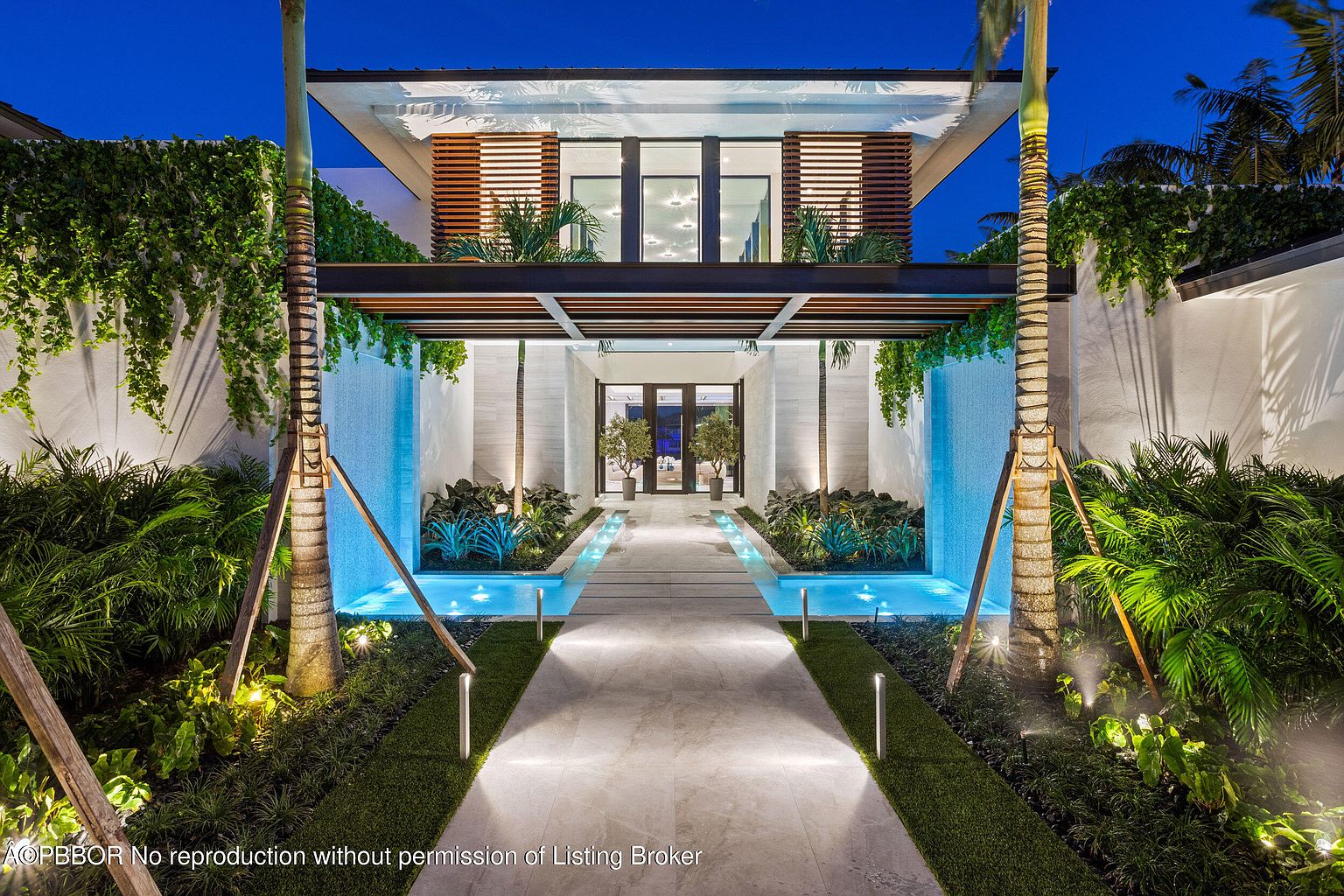 This is a stunning entryway to a modern home, featuring a long, illuminated walkway leading to the front door. The walkway is flanked by water features, lush greenery, and palm trees, creating a serene and inviting atmosphere. The architectural design incorporates clean lines, natural materials, and ambient lighting, enhancing the property's curb appeal and creating a luxurious first impression.