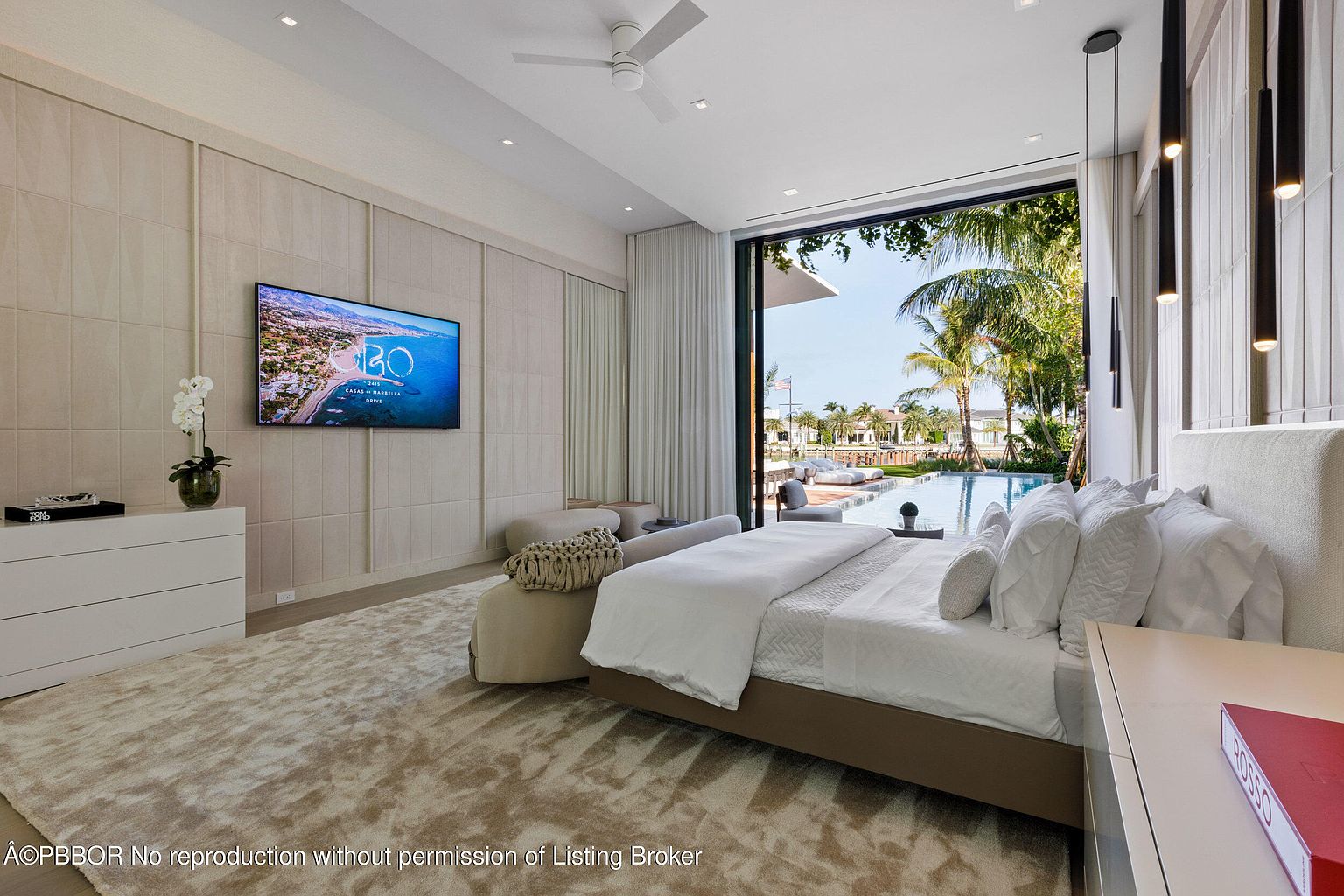 This is a luxurious primary bedroom featuring a king-size bed with white linens, a textured wall with a large flat-screen TV, and floor-to-ceiling windows offering a view of a pool and palm trees. The room is decorated in neutral tones with modern lighting fixtures, creating a serene and upscale atmosphere. The overall impression is one of comfort and sophistication.