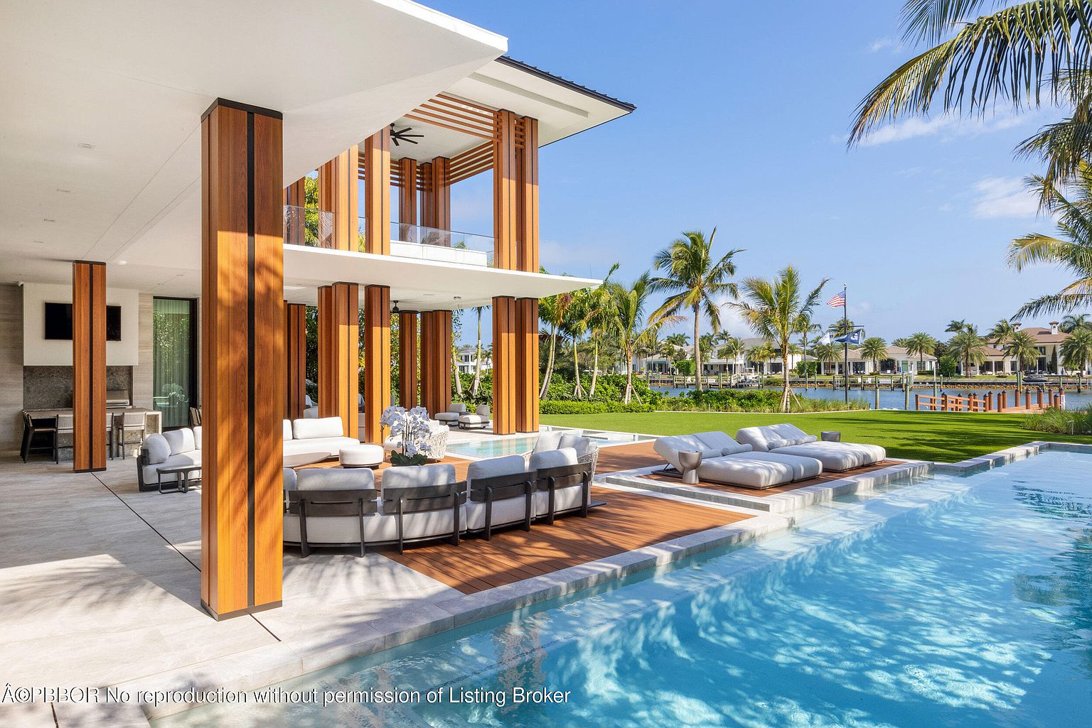 This exterior shot showcases a luxurious pool area with modern architectural design. The pool features clear blue water and is surrounded by a patio with stylish outdoor furniture, including lounge chairs and a curved sofa. The structure of the house includes wooden support beams and an open-air design, with a view of a well-manicured lawn and waterfront property in the background.